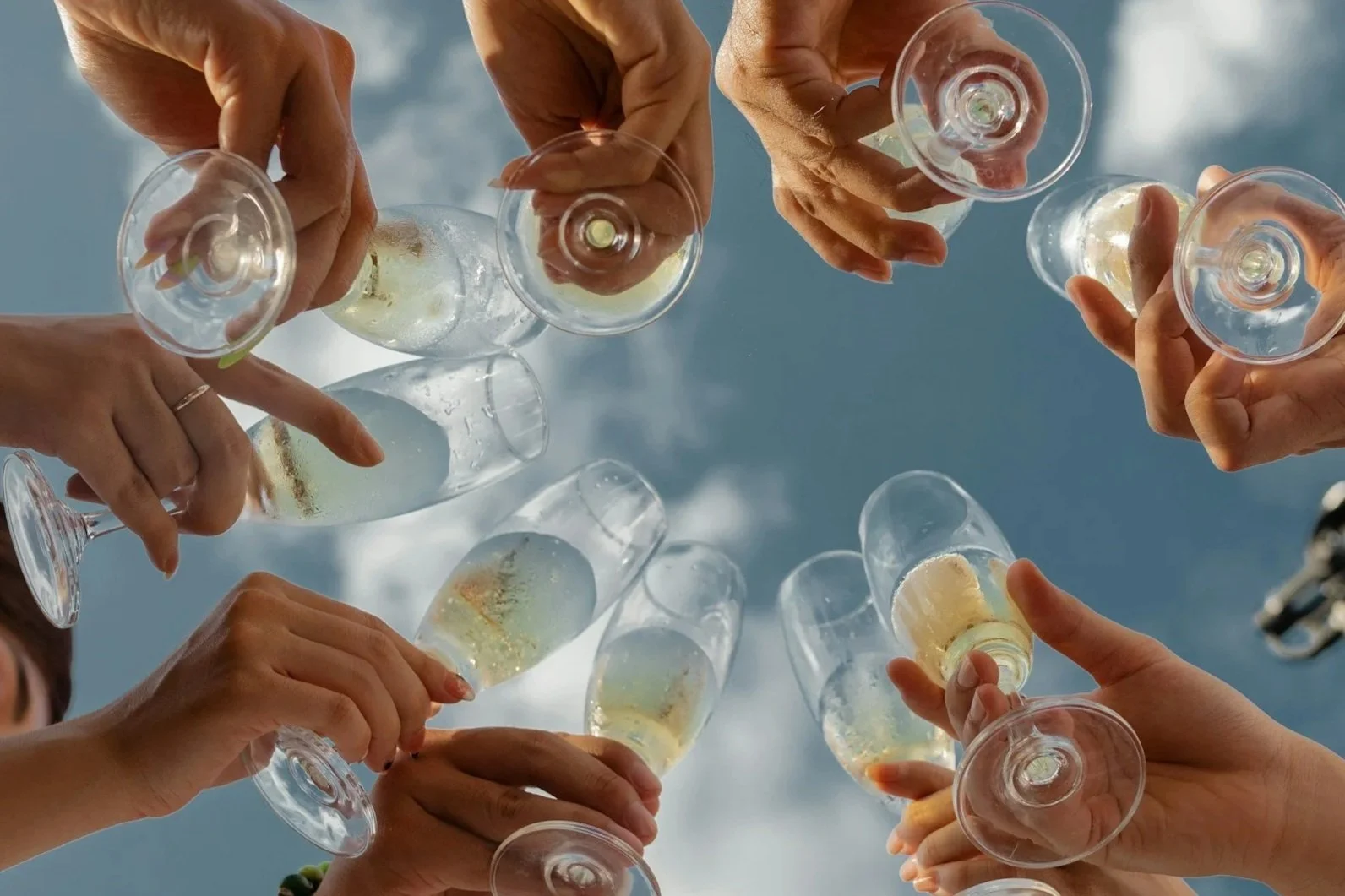 Group of people holding champagne flutes filled with sparkling wine, raised in a toast outdoors against a blue sky. Celebration travel