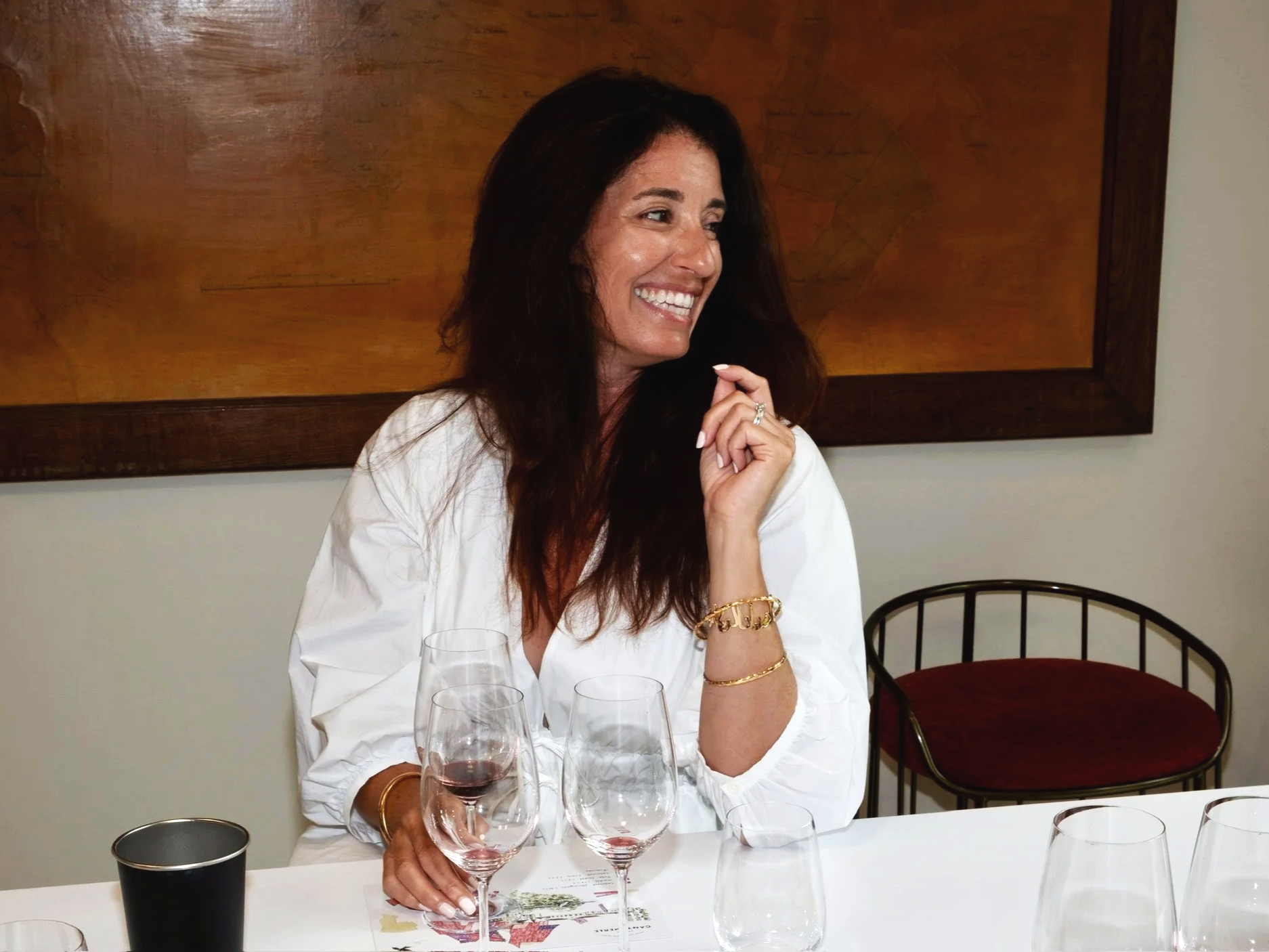A woman with dark brown hair laughing and smiling, sitting at a table with wine glasses, a black cup, and a piece of paper, in a room with a large wooden framed map or artwork on the wall behind her.