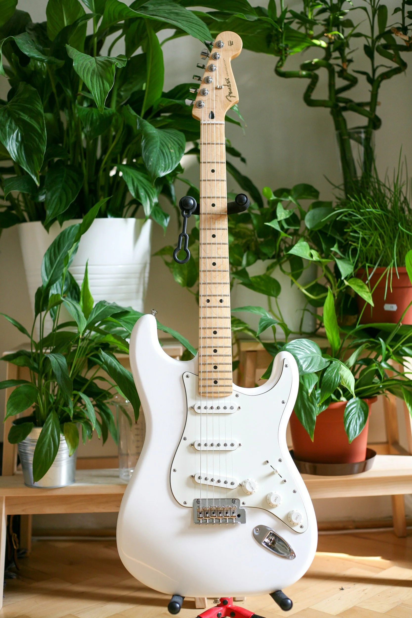 A white electric guitar is standing on a wooden floor in front of a wooden table with potted green plants. The guitar has a maple neck and headstock with a Fender logo, and is equipped with pickups, control knobs, and a chrome bridge.