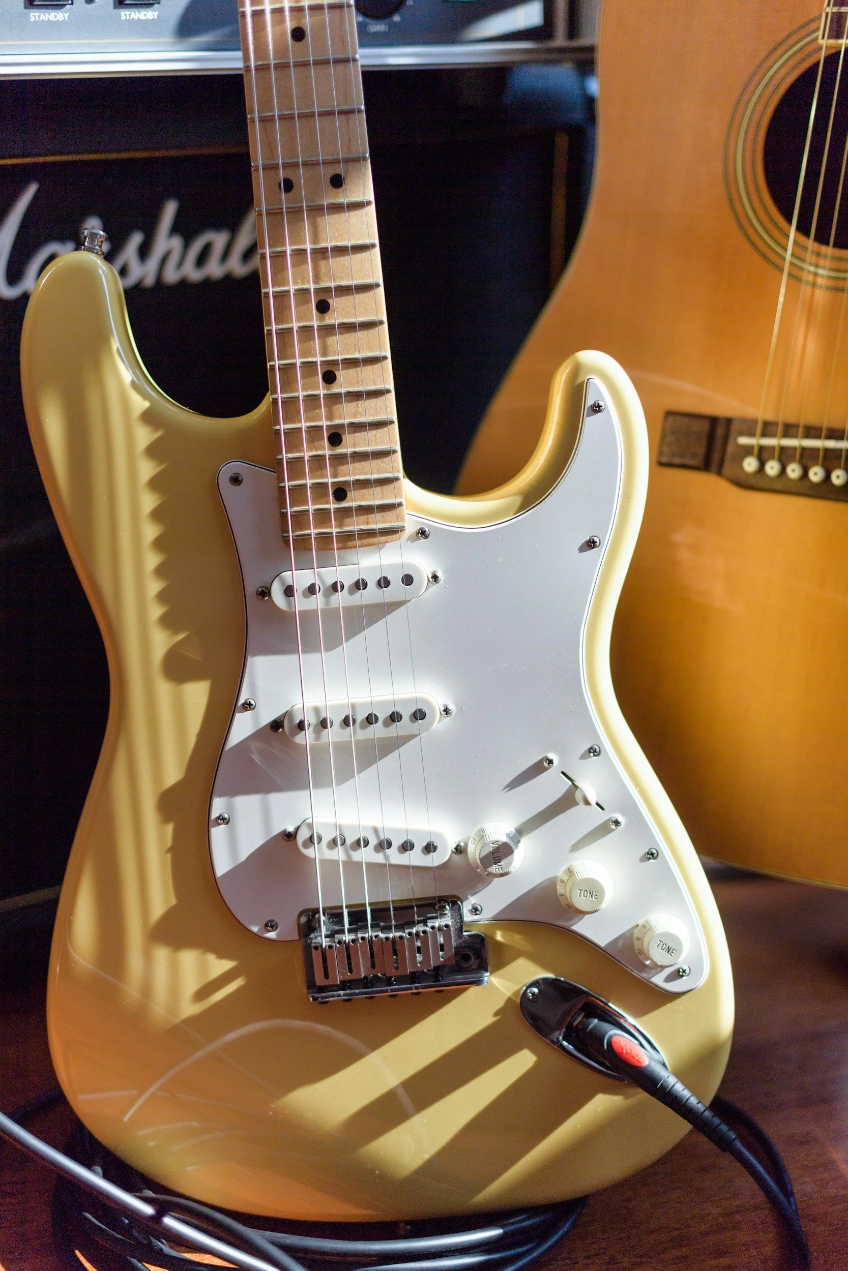 Close-up of a yellow electric guitar with a white pickguard, connected to an amplifier with a black cable. A wooden acoustic guitar is visible in the background.