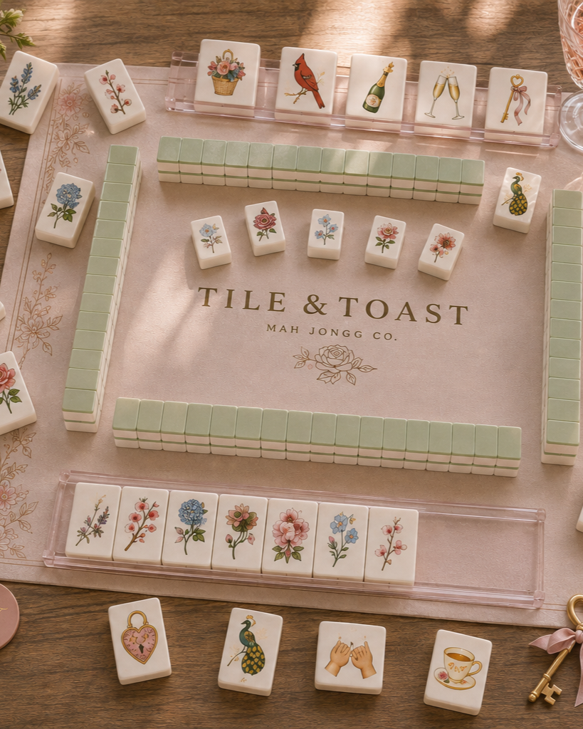 Close-up of a mahjong game set featuring floral-themed tiles and decorative tiles with images of a basket of flowers, a cardinal bird, champagne, and a key, on a table with a pink mat.
