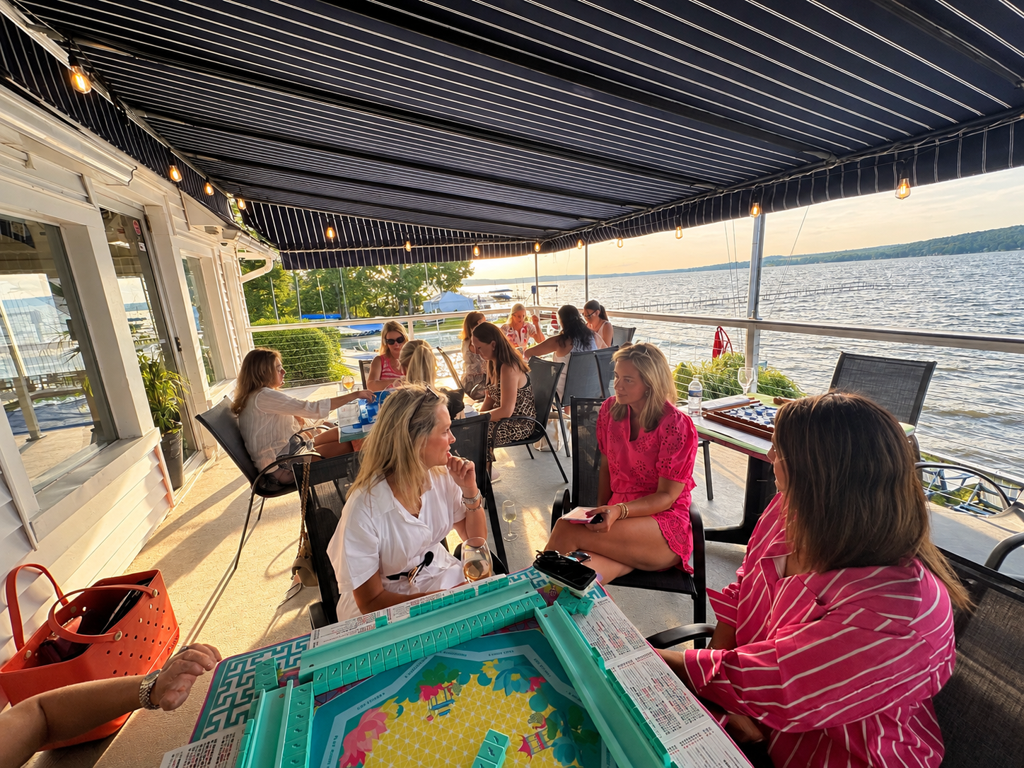 Group of women sitting on a boat deck overlooking the water at sunset, playing American Mah Jongg and socializing.