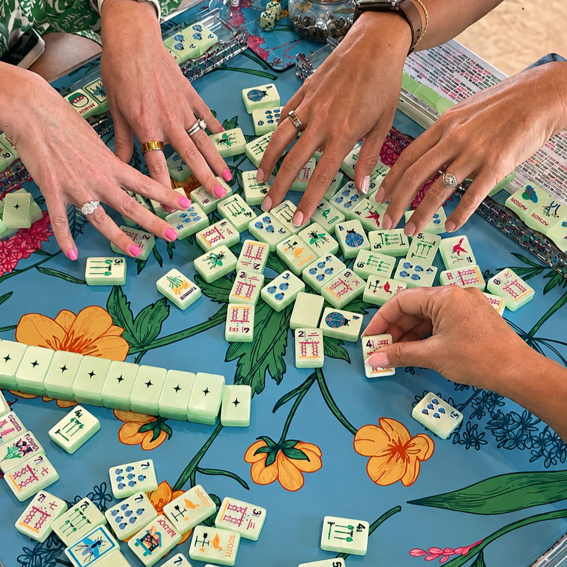 Three people playing a game of Mahjong at a table covered with a blue floral tablecloth, with Mahjong tiles scattered across the table.
