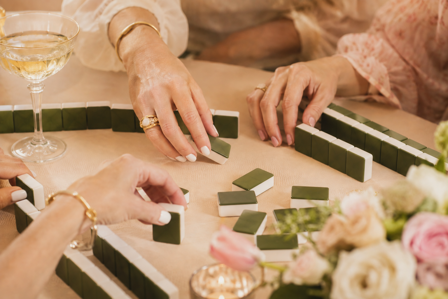 Close-up of women playing mahjong at a table with a beige tablecloth, a glass of white wine, and a bouquet of pink and white flowers.