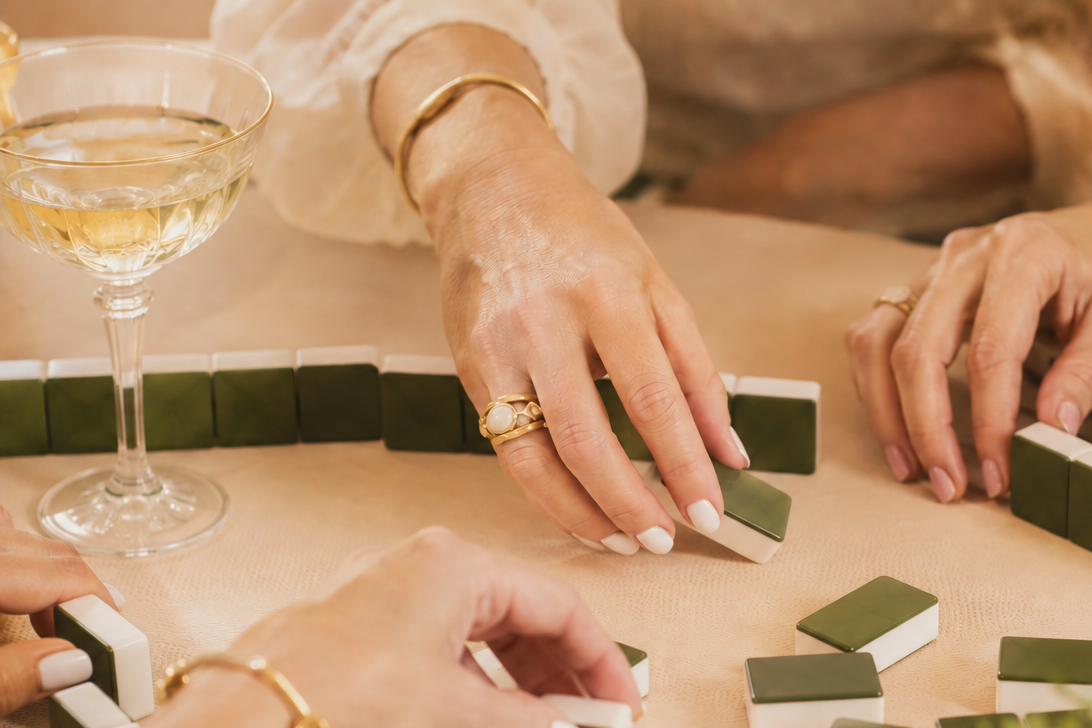 Close-up of hands with jewelry playing Mahjong, with a glass of champagne on a table.