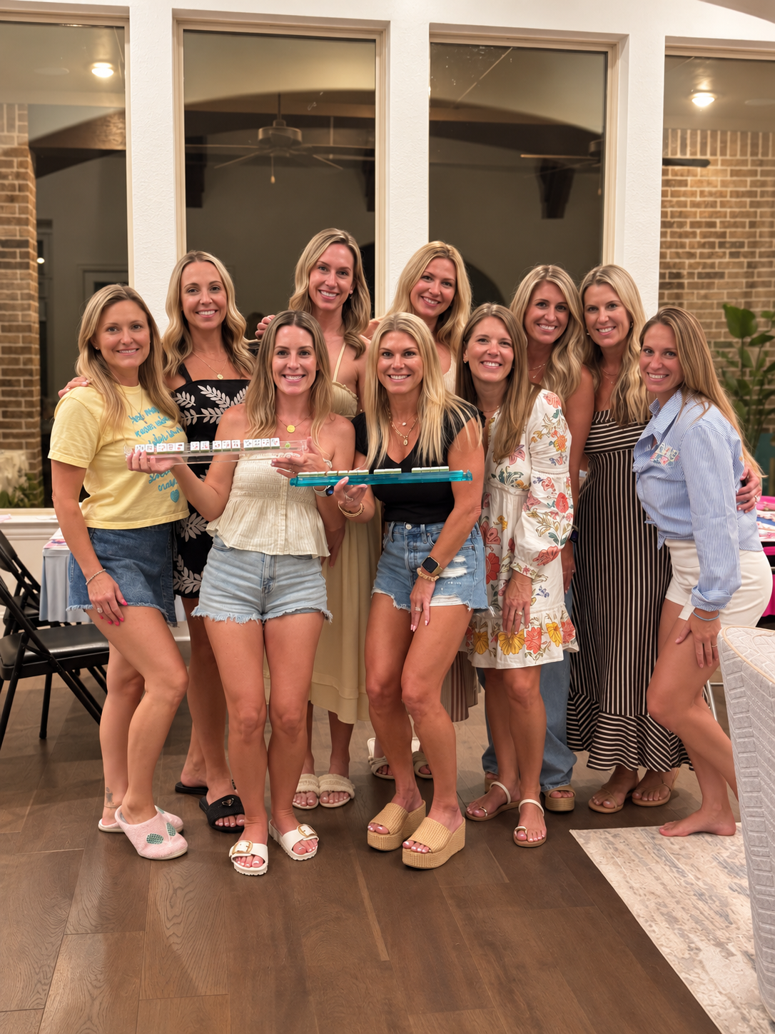 A group of eleven women gathered indoors at a celebration, with some holding drinks and party accessories. They are smiling and standing close together in front of large windows, with a cozy, well-lit interior behind them.