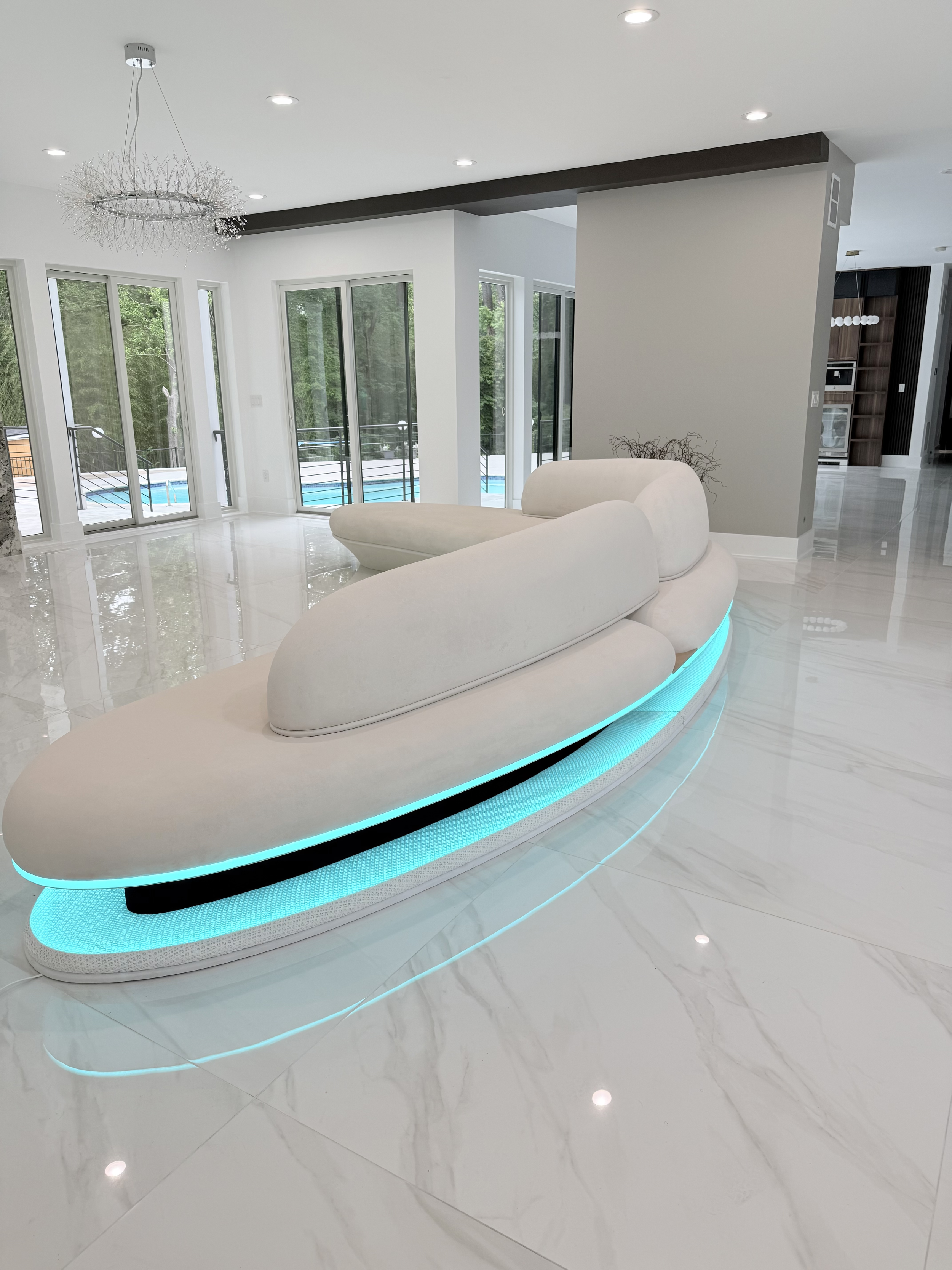 Modern living room with white luxury curved sofa with glowing blue base, large windows overlooking trees and pool, and white marble flooring.