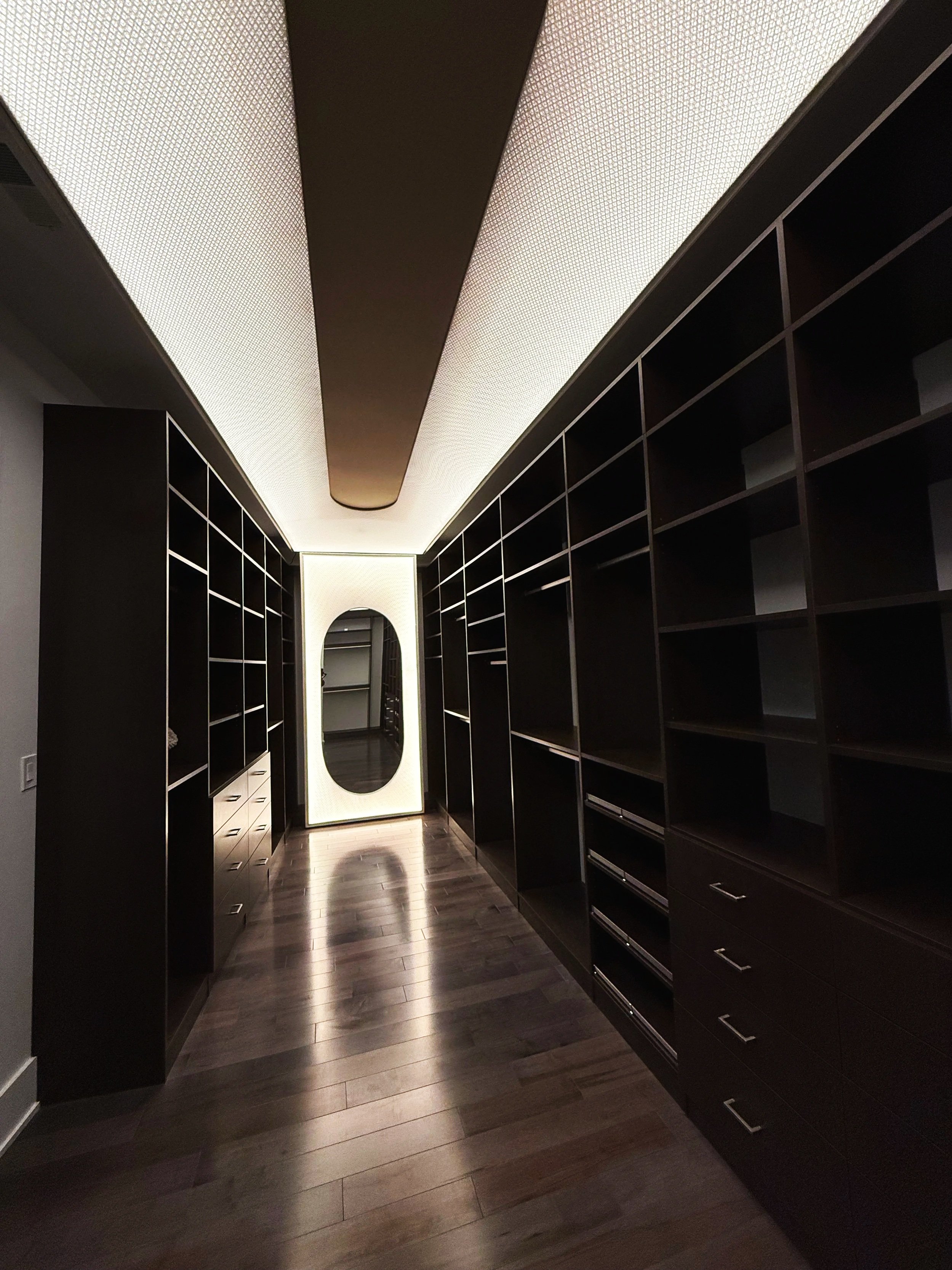 A walk-in closet with dark wooden shelves and drawers on both sides, a full-length mirror at the end, and a dark wood floor.