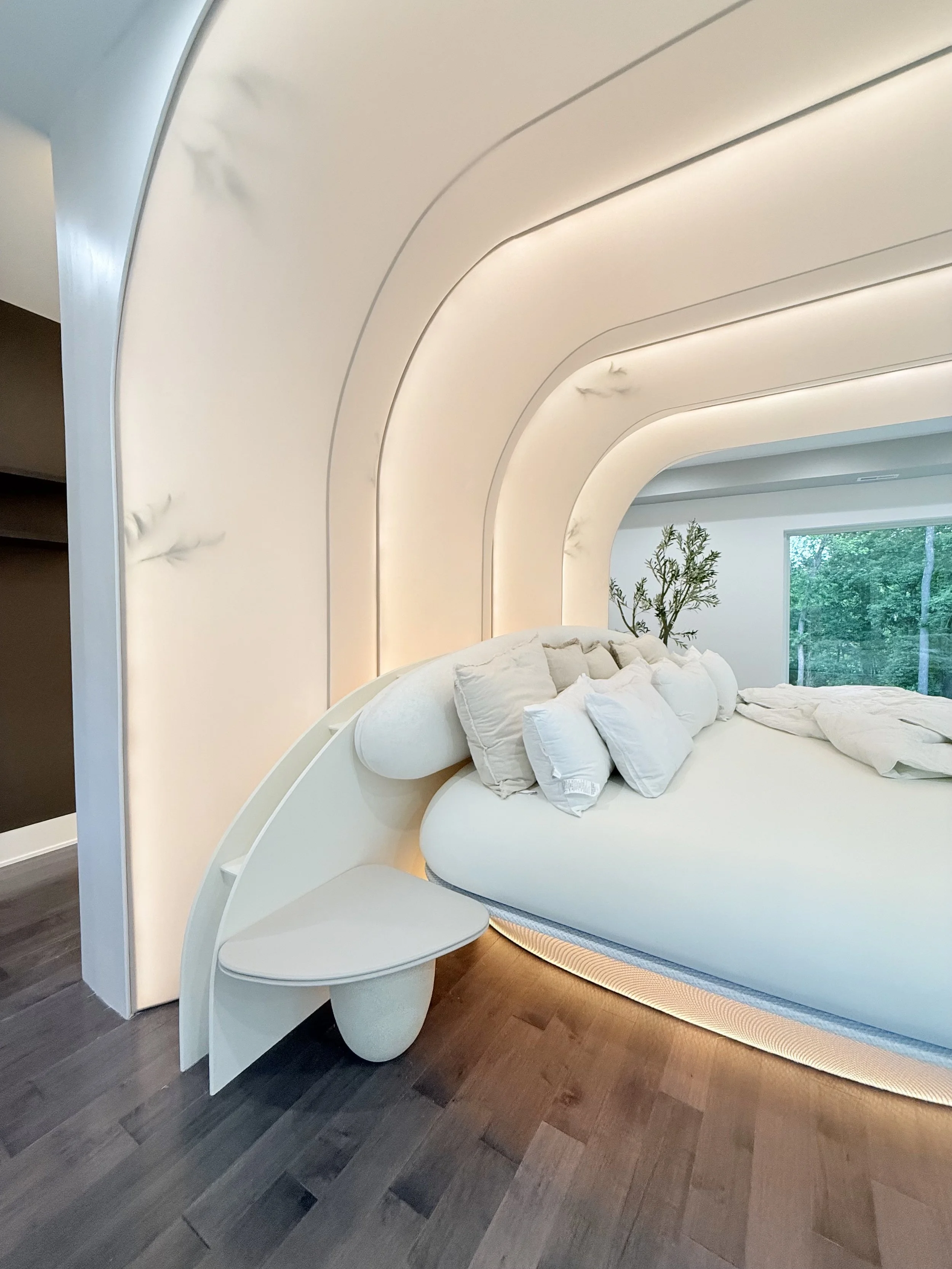 Modern bedroom with curved white headboard, white bed linens and pillows, a small round bench at the foot of the bed, and a window showing green trees outside.