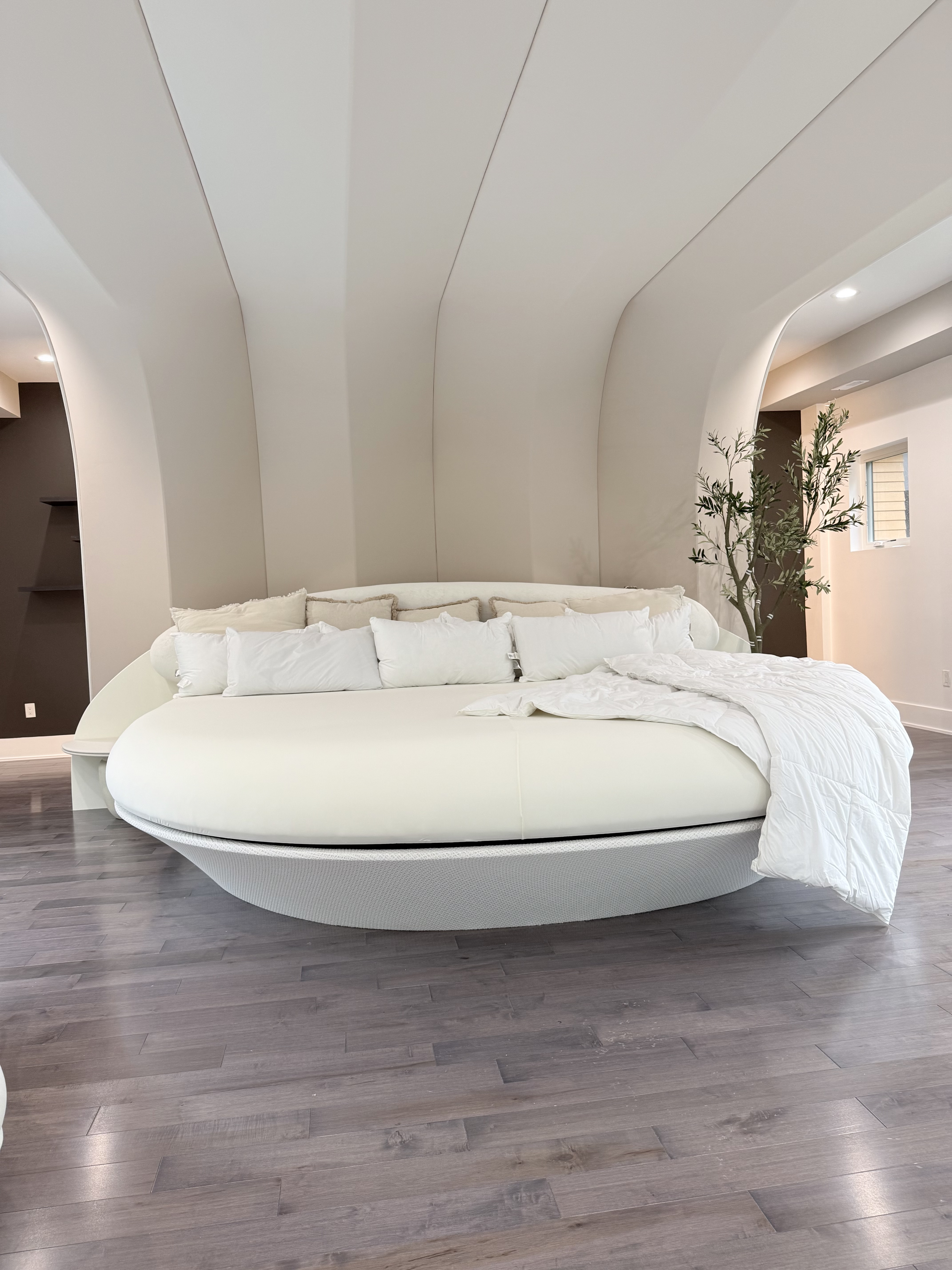 Modern bedroom with a curved white bed, white bedding, throw pillows, a tall green plant, light gray hardwood floors, and minimalist wall design.