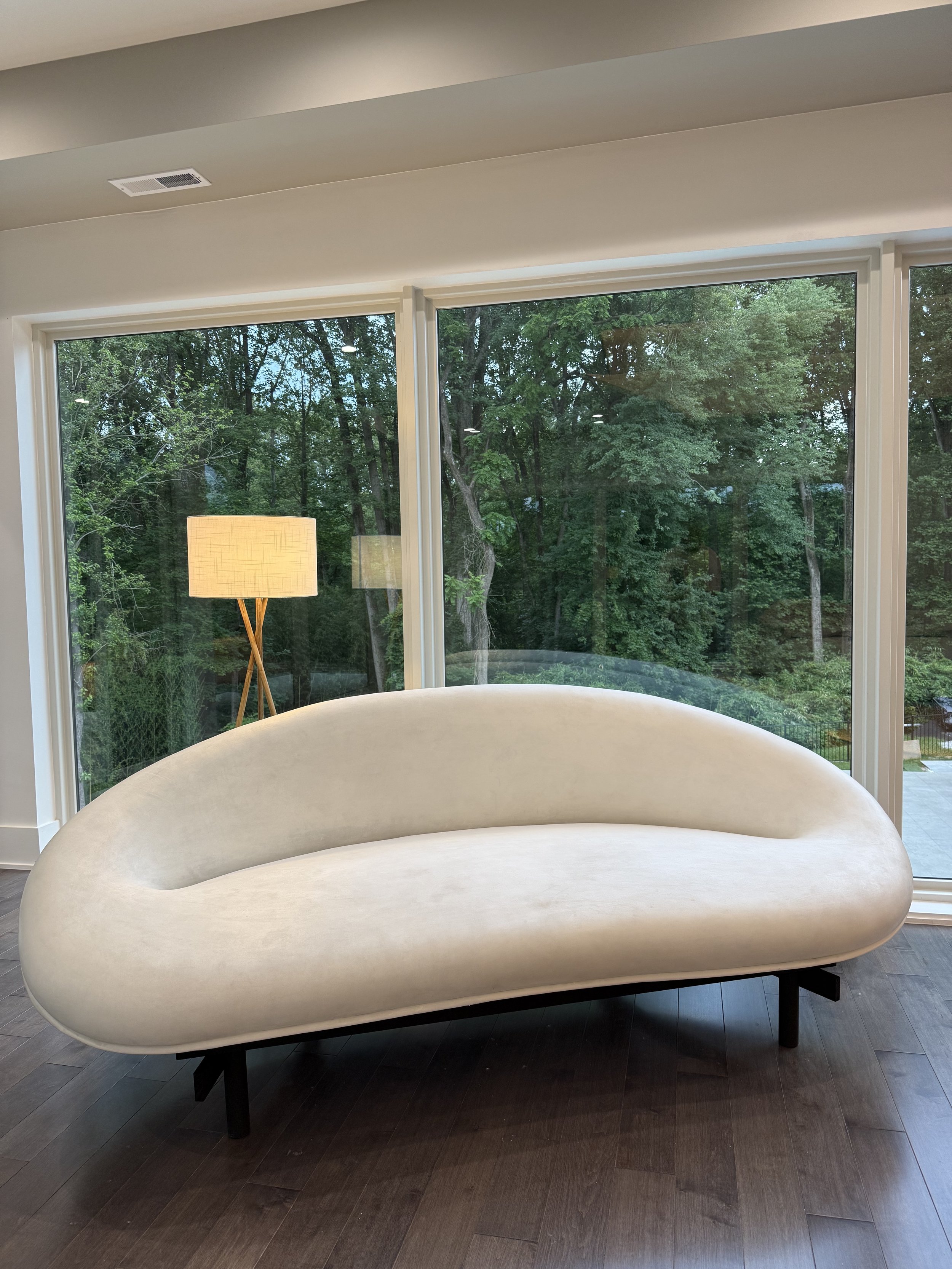Modern indoor space with a large curvy beige sofa on dark wooden floors, glass windows showing green trees outside, and a tall floor lamp with a beige shade.