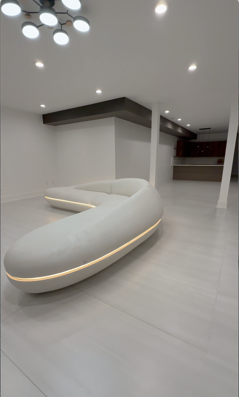 Modern living room with curved white sofa with LED lighting, white walls, light-colored flooring, and modern ceiling lights.