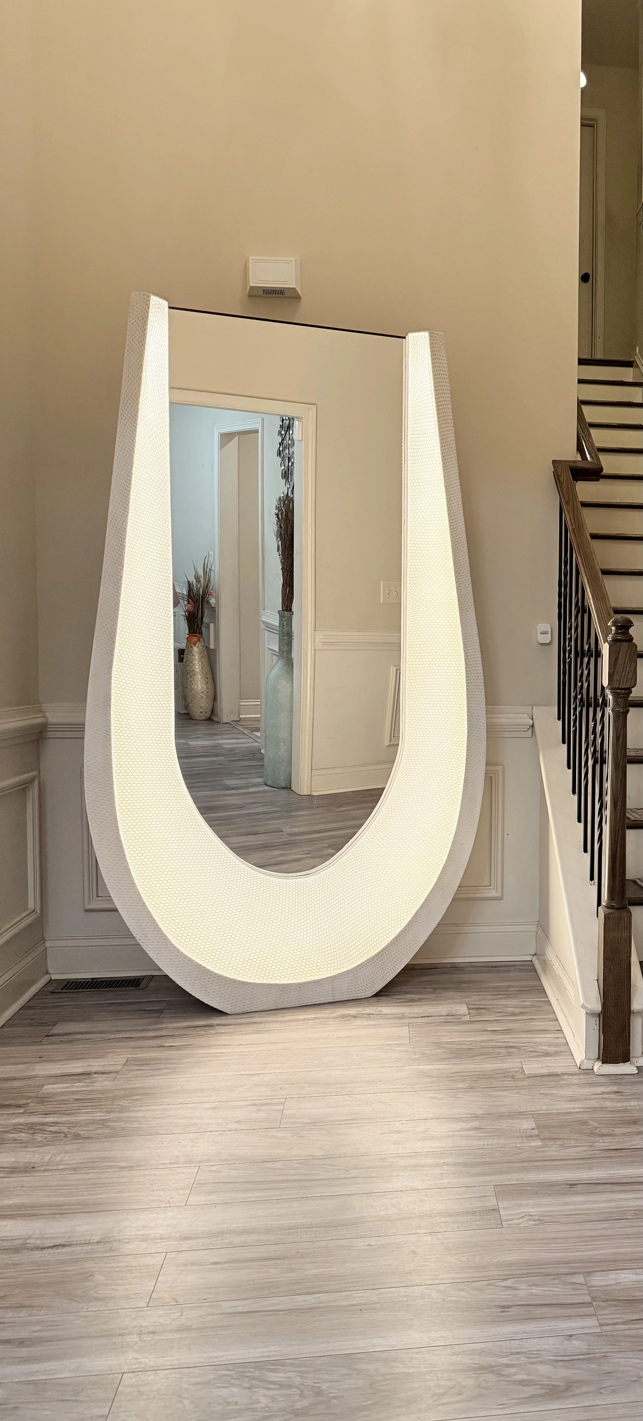 A large illuminated U-shaped mirror leaning against a beige wall in a home interior. Futuristic mirror