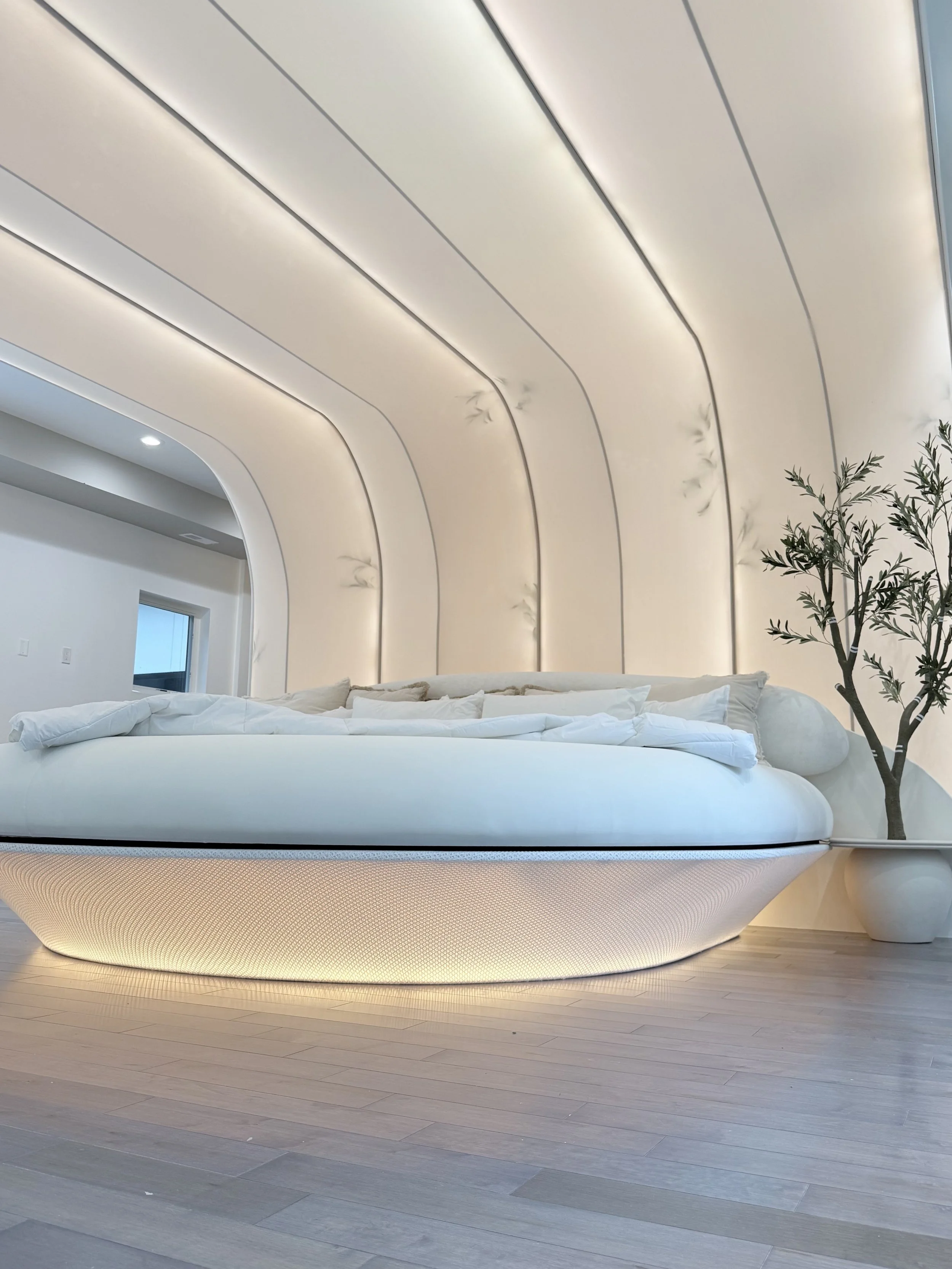 Minimalist modern room with a curved white bed, soft beige padded wall panels ceiling, wooden floor, and a potted plant.