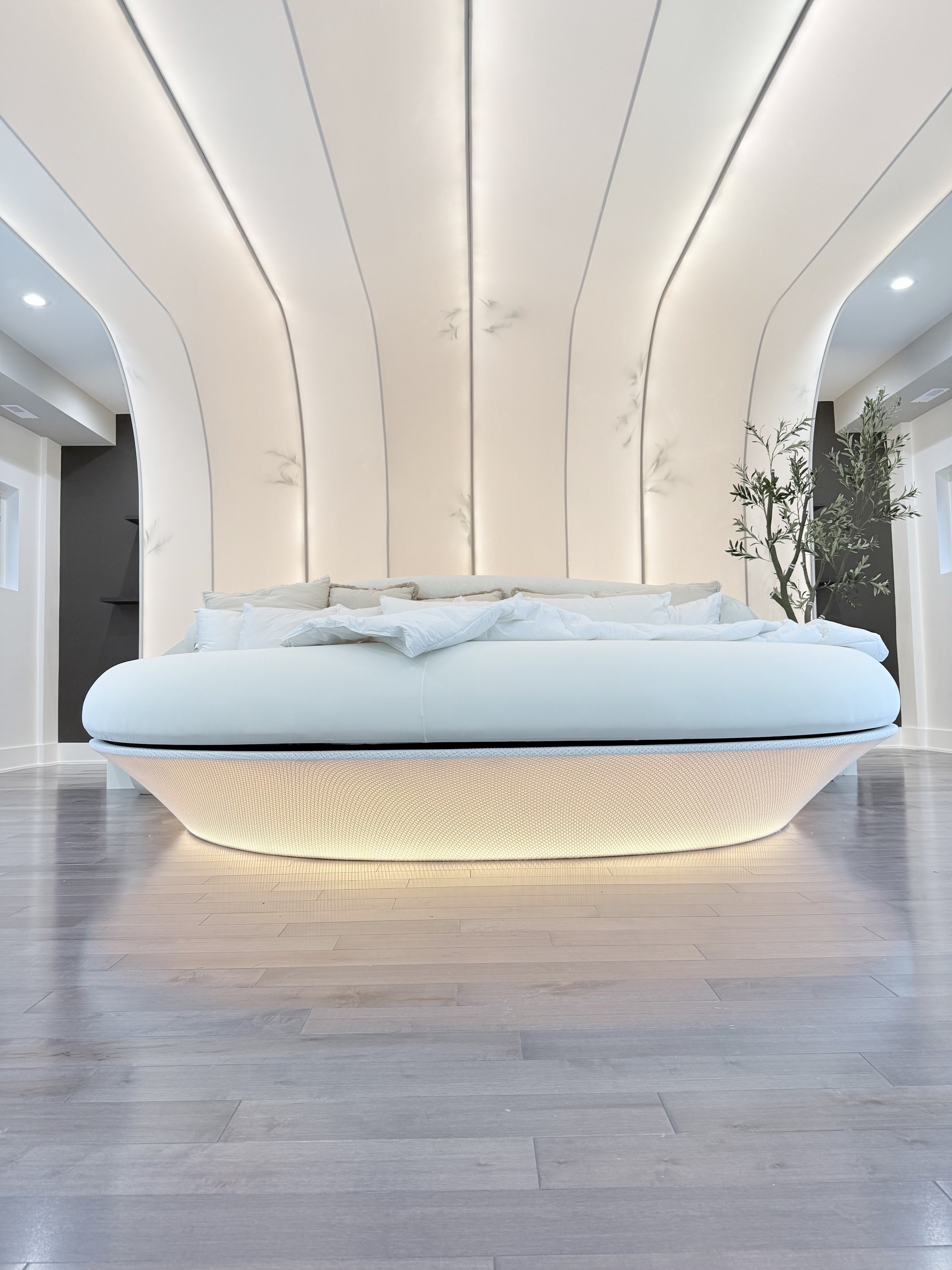 Modern bedroom with a cushioned bed with white bedding, behind a curved white headboard, and a tree with green leaves on the right side, set against a white and gray wall with decorative designs.