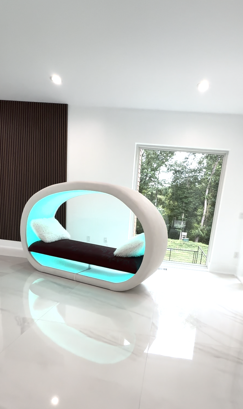 Modern luxury white oval-shaped lounge pod/chair with blue lighting, placed in a bright room near large window showing greenery outside, with white pillows on the seat.