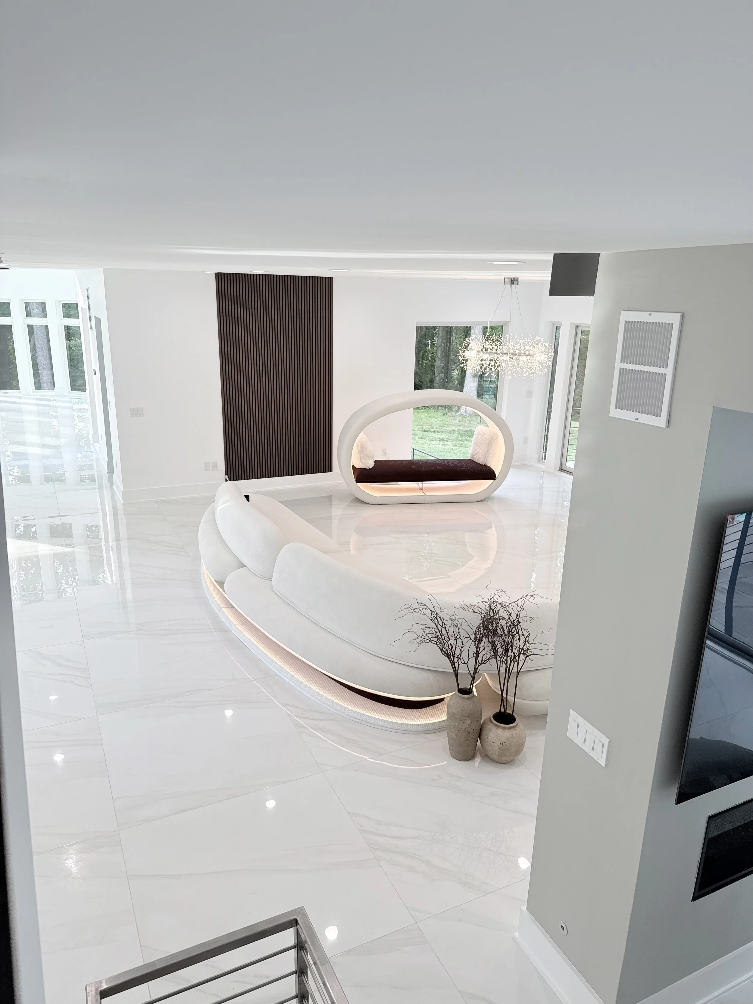 Modern, minimalist interior with white walls and glossy white tile floors. A unique oval-shaped bench with a dark wooden top and white curved sides is located in the center. Two beige vases with dried branches sit on the floor nearby. Large windows show greenery outside, and a contemporary chandelier hangs from the ceiling.