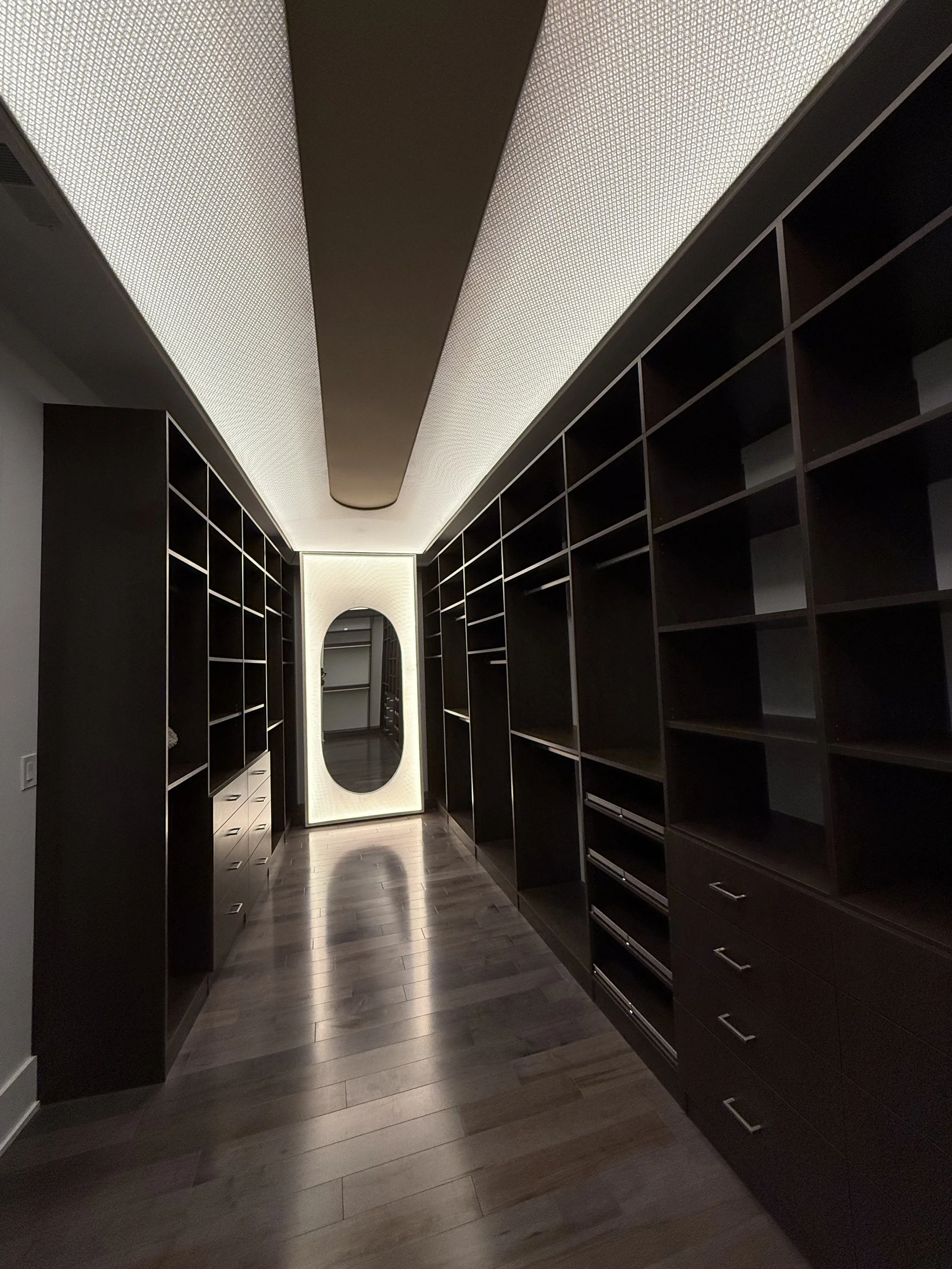 A corridor with built-in dark wooden shelves on both sides, leading to a white backlit oval mirror at the end, and dark wood flooring.