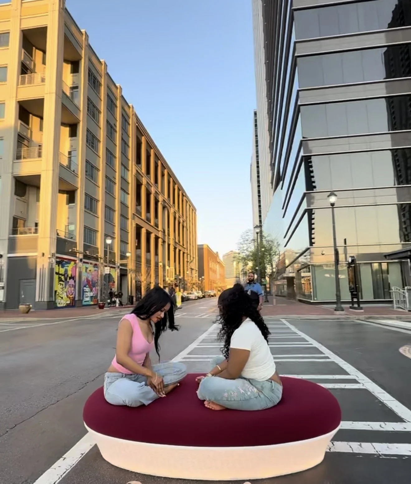 Two women sitting cross-legged on a red luxury ottoman bench with lighting in the middle of a city street, with tall modern buildings on either side.