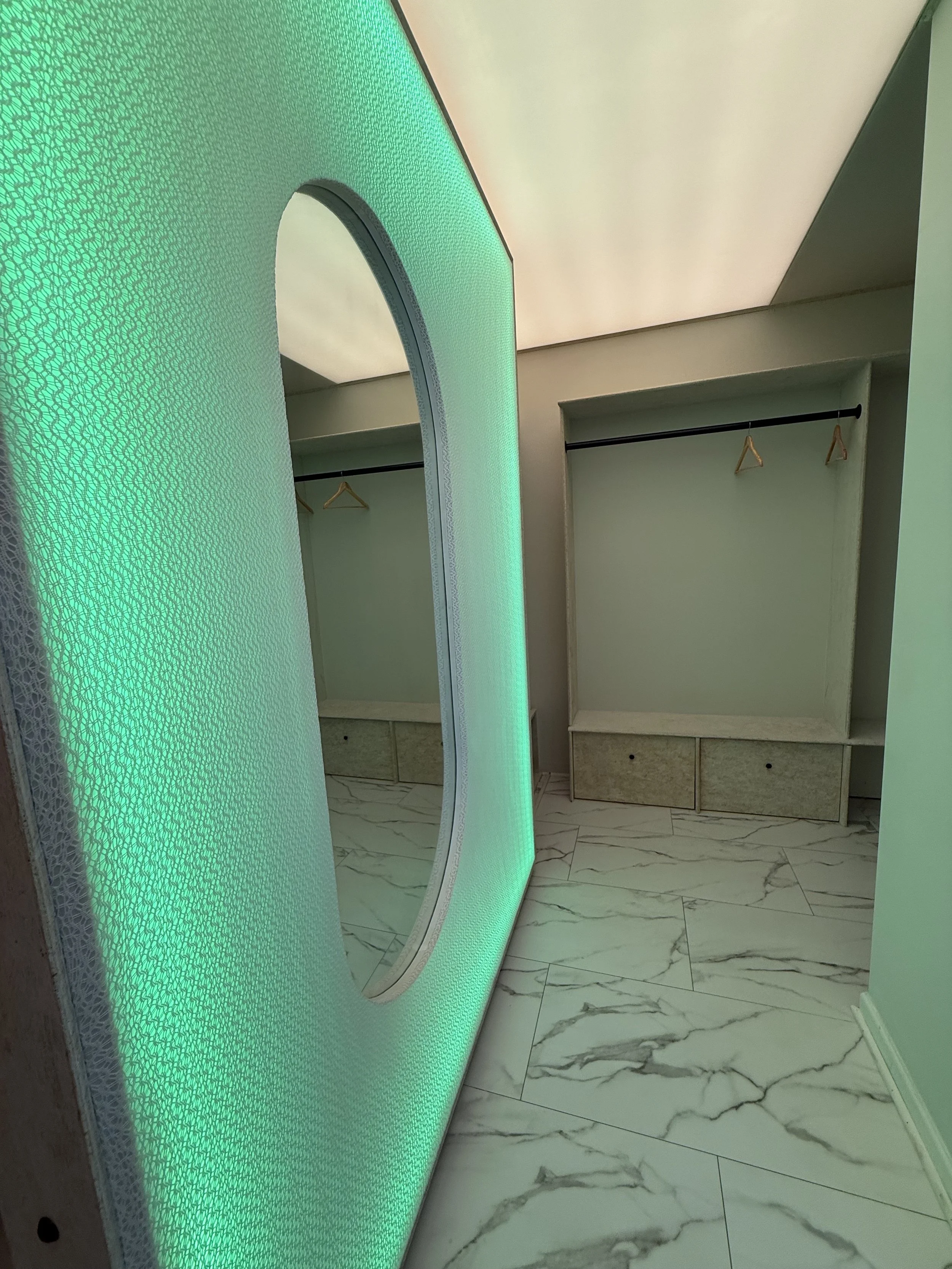 A modern, stylish closet with green and white ambient lighting, a large oval luxury futuristic mirror with lighting. 