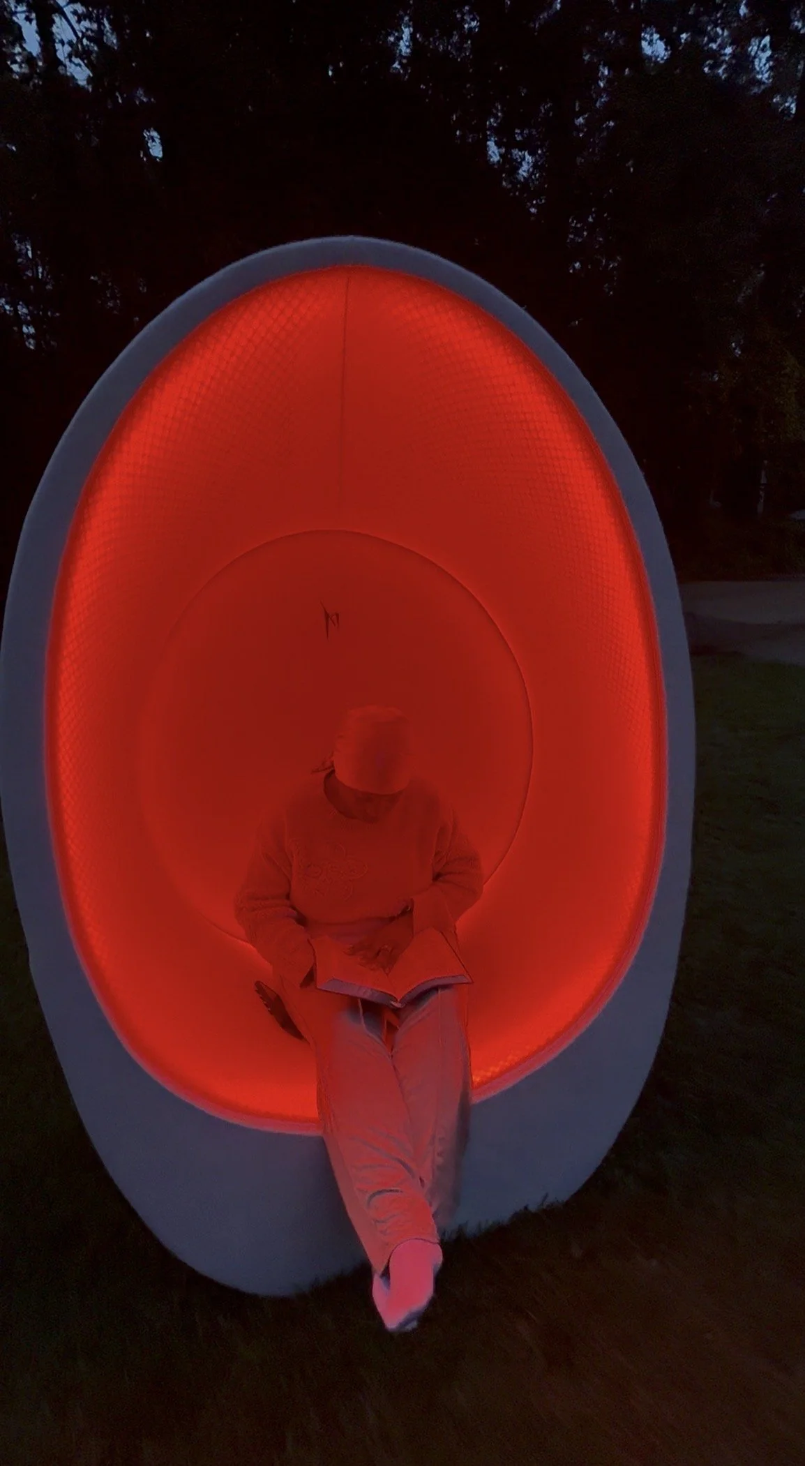 Person sitting inside a red-lit pod-shaped structure at night, reading a book.