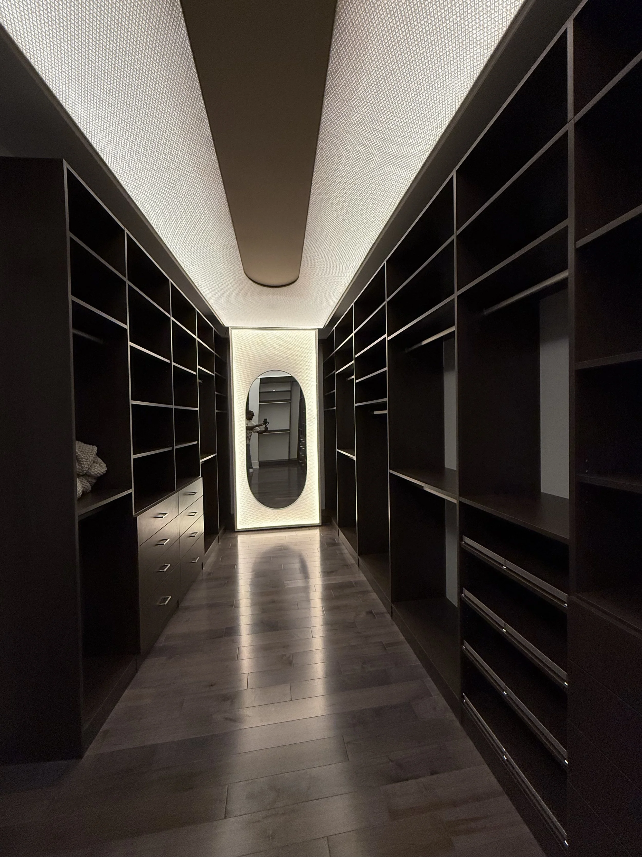 Modern walk-in closet with futuristic ceiling light , dark wood cabinets and shelves, a full-length luxury mirror, and wooden flooring.