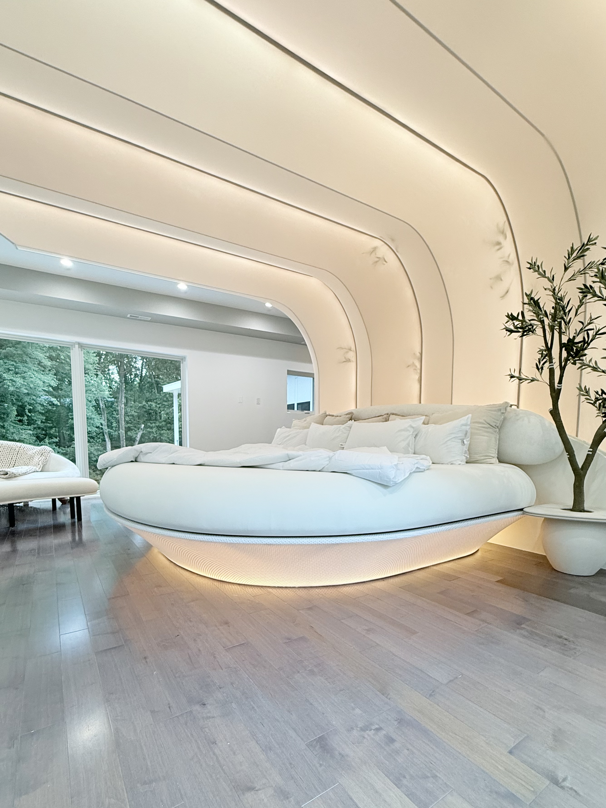 A unique futuristic bedroom with curved lit ceiling design, large luxury and unique bed with white bedding and pillows, potted plant, and large window with trees outside.