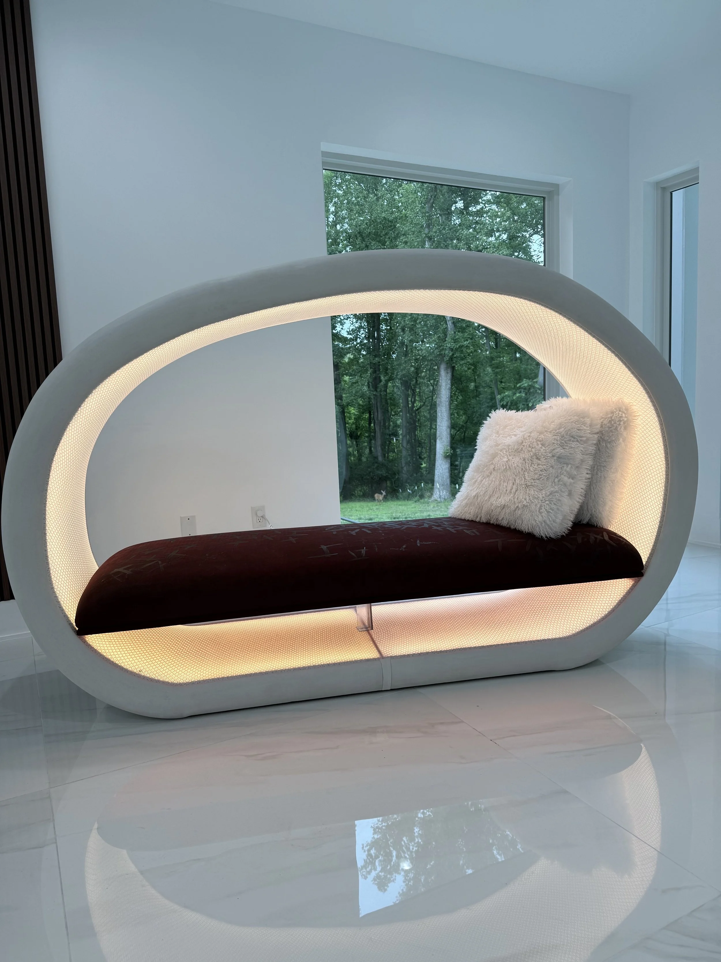 Modern luxury white oval-shaped lounge pod/chair with lighting, placed in a bright room near large window showing greenery outside, with white pillows on the seat.