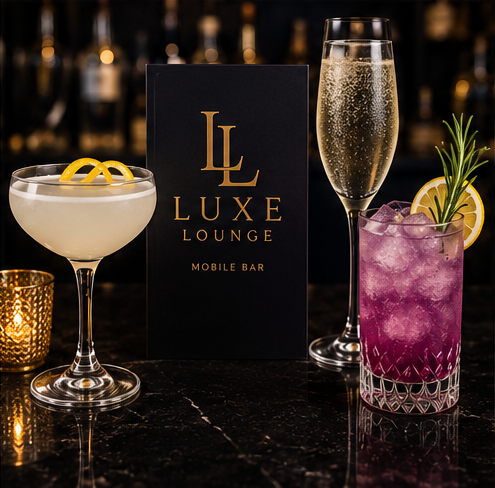 Two cocktails on a bar counter with a Luxe Lounge sign in the background. The cocktail on the left is a pale yellow with lemon twists, the middle is a tall glass of sparkling wine, and the right is a pinkish-purple drink with lemon, rosemary, and ice.