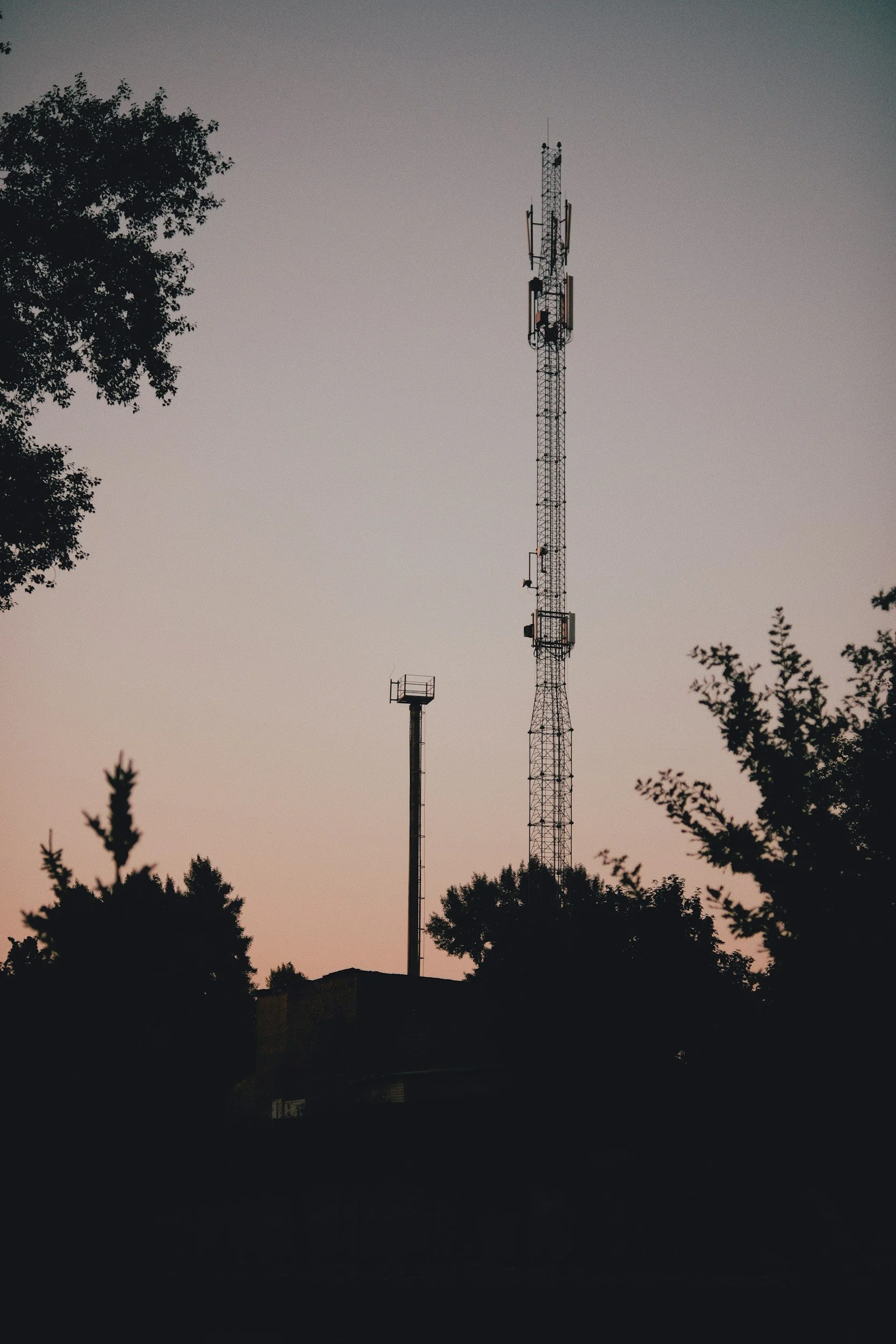 A tall communication tower with various antennas and dishes, silhouetted against a dusky sky, with trees in the foreground.