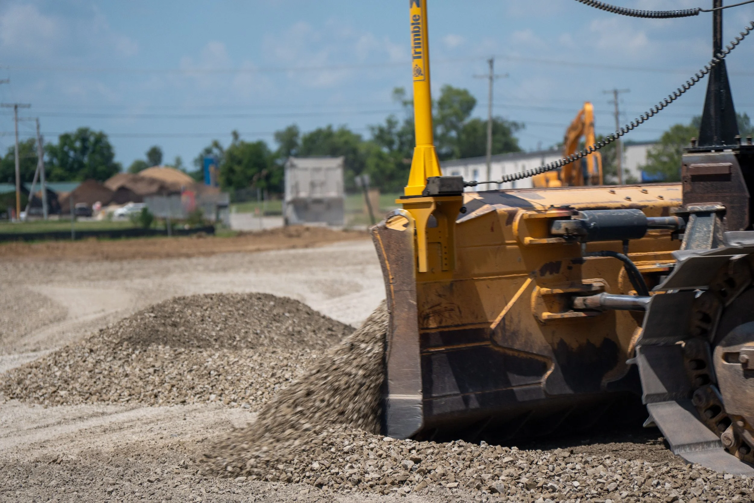 Construction vehicle spreading gravel or dirt on a construction site with houses, trees, and power lines in the background.