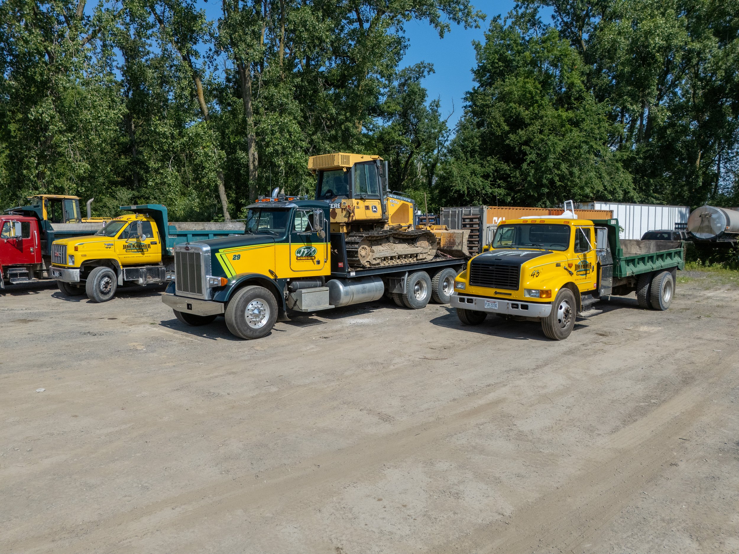A group of trucks parked on a dirt lot, with a yellow excavator on a flatbed truck, surrounded by tall trees and a clear blue sky.