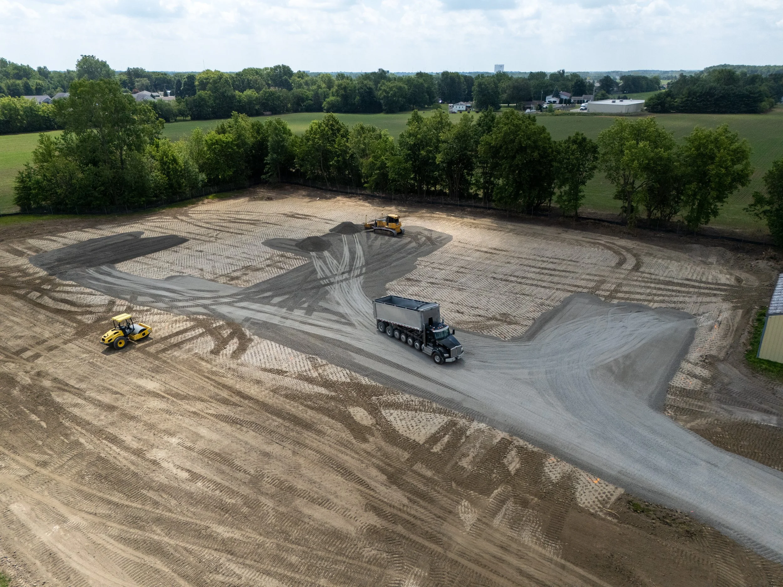 Construction site with cleared land, construction vehicles, and dirt piles, surrounded by green trees and rural scenery.