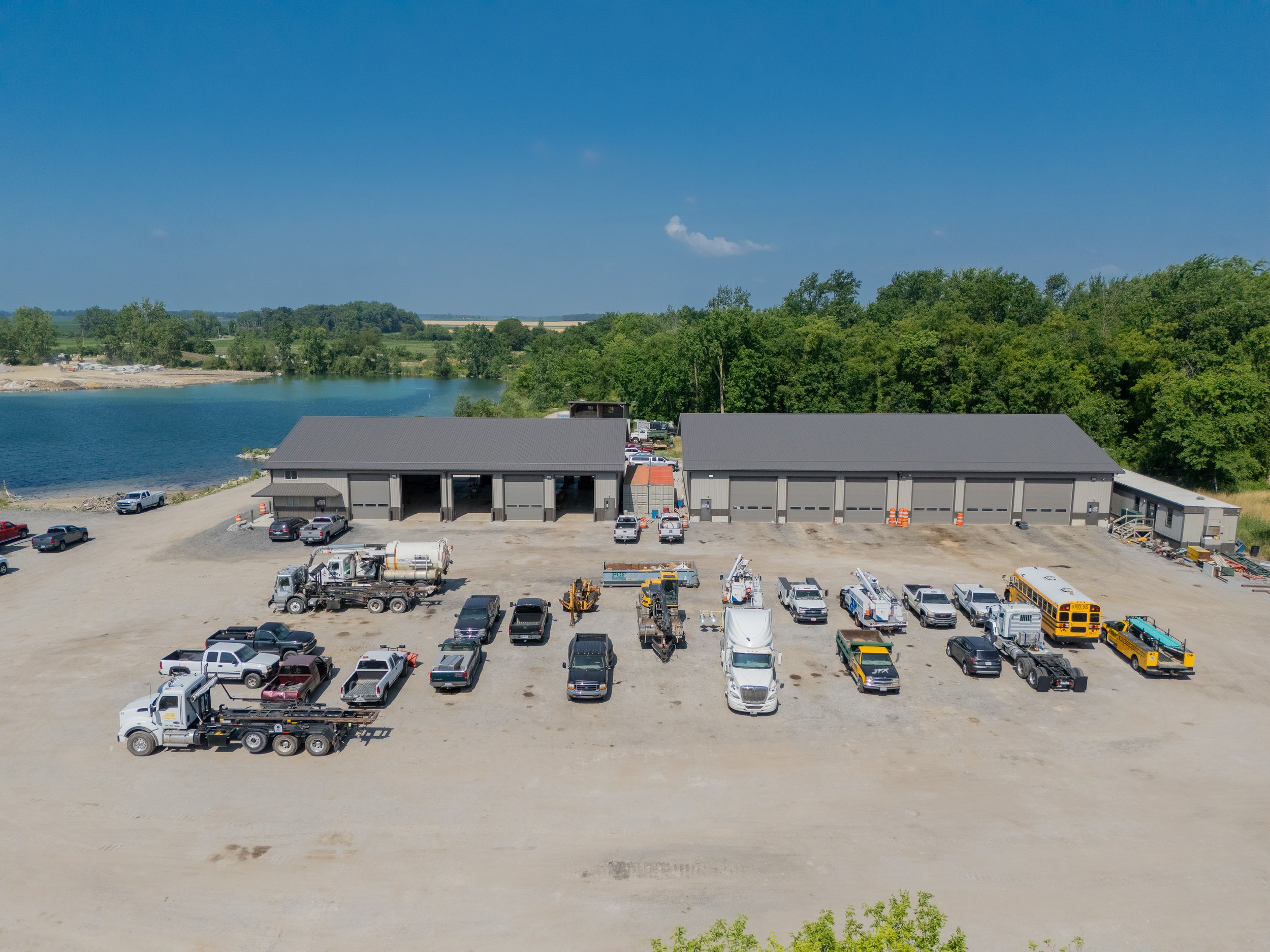 A parking lot with various vehicles in front of large buildings under a blue sky with some clouds, and a landscape of trees and fields.