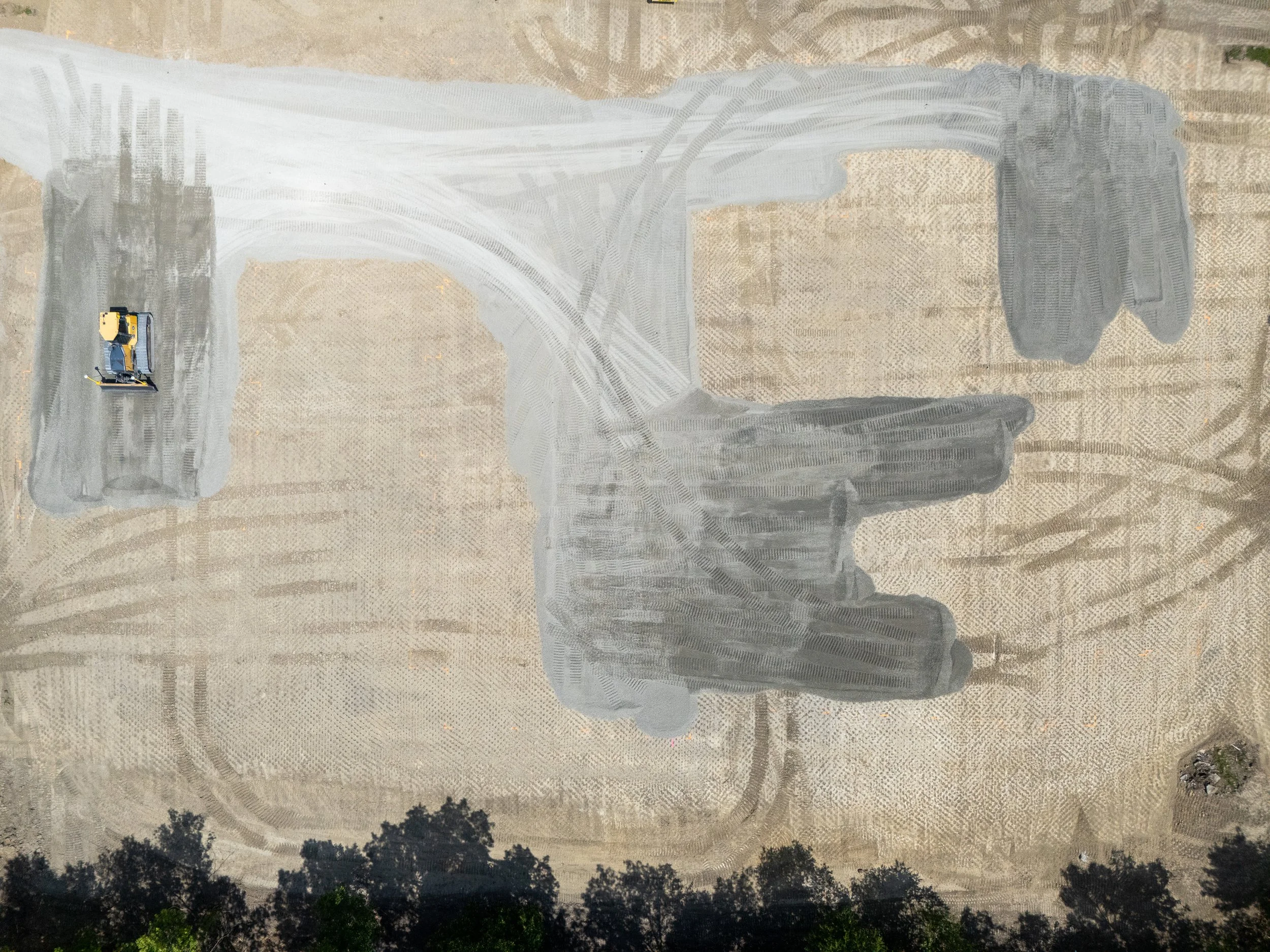 A construction worker in a yellow and black safety suit with a yellow hard hat is operating a steamroller on a large area of dusty land. The land appears to be in the process of being leveled or prepared, with visible tire tracks and some trees in th