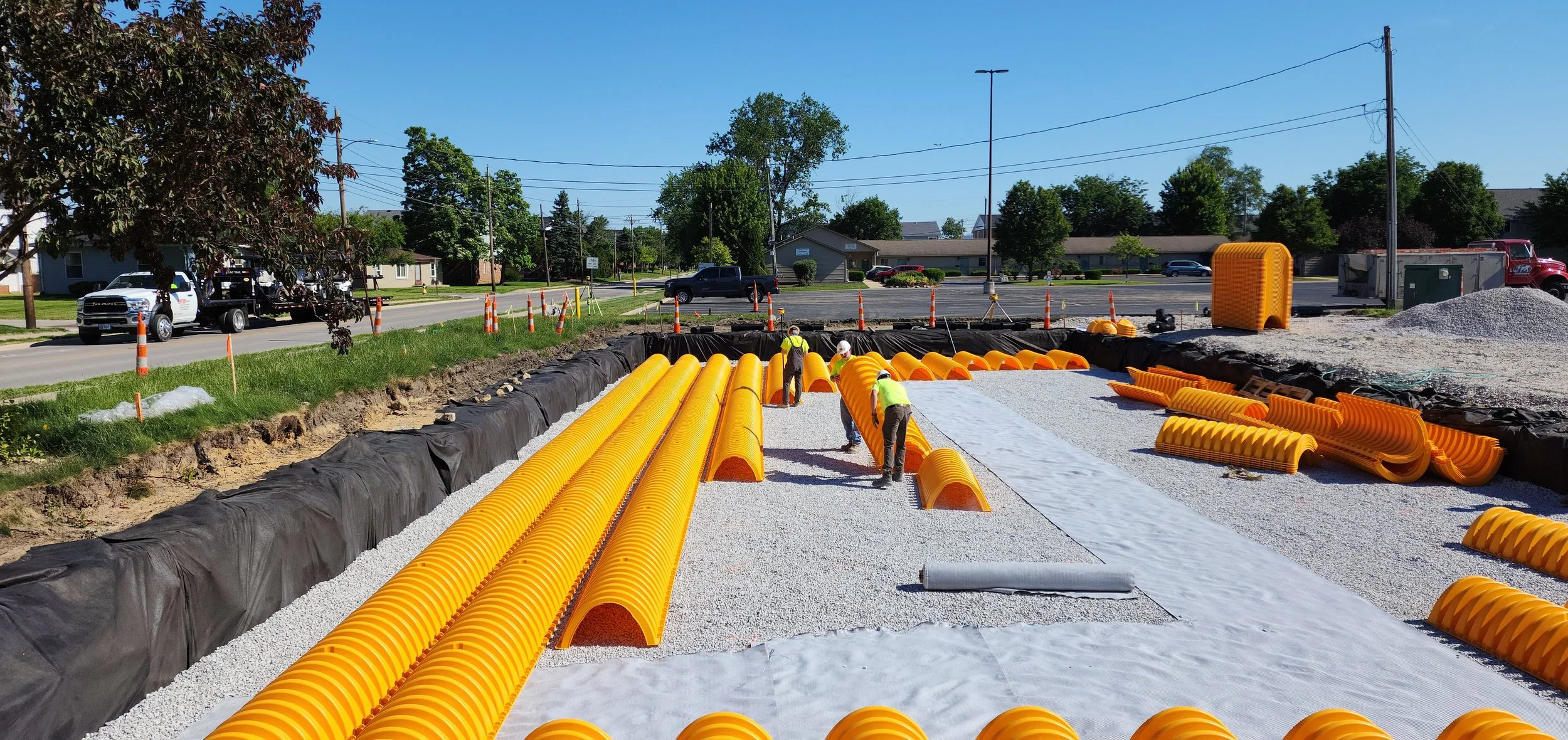 Construction workers installing orange protective culverts in a construction site along a sidewalk, with trees, houses, and cars in the background on a sunny day.