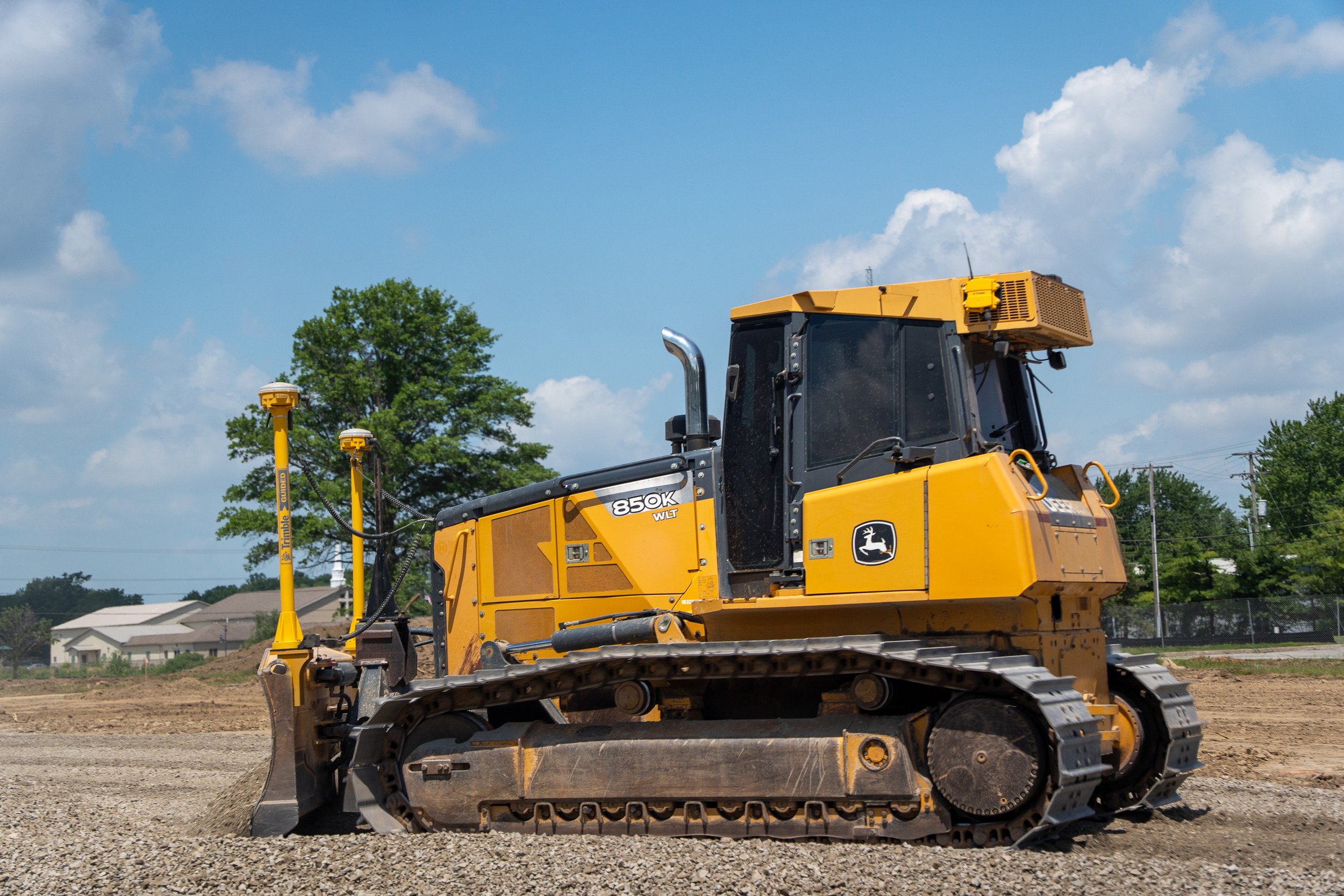 A yellow John Deere bulldozer working on a construction site with trees and houses in the background under a partly cloudy sky.