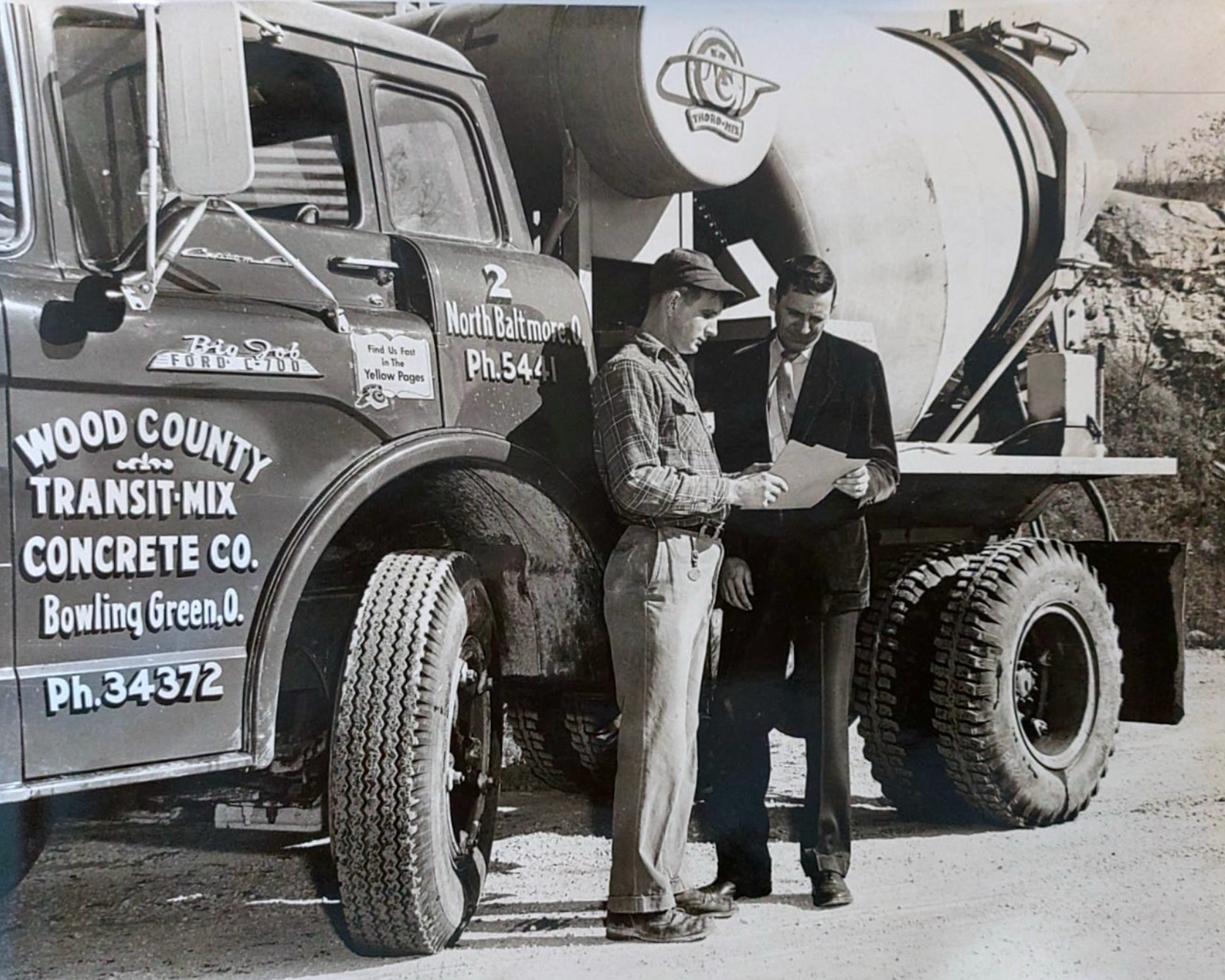 Two men stand beside a vintage service truck with large tires. One man is dressed in a checkered shirt and light-colored pants, the other in a dark suit. They are looking at a piece of paper. The truck has signs indicating it is associated with a transit and concrete company in Bowling Green, Ohio.