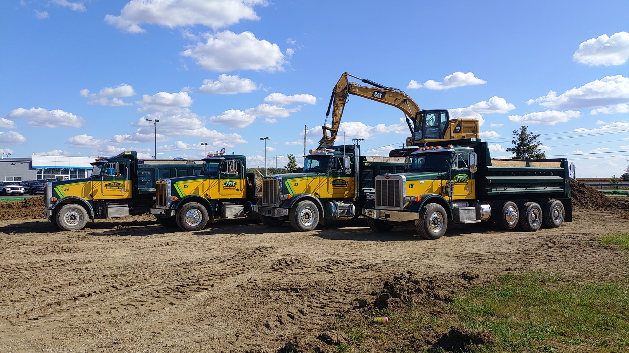 Four large construction trucks parked on a dirt lot with a bulldozer on top of two of the trucks. The background includes a blue sky with white clouds and a commercial building with cars parked outside.