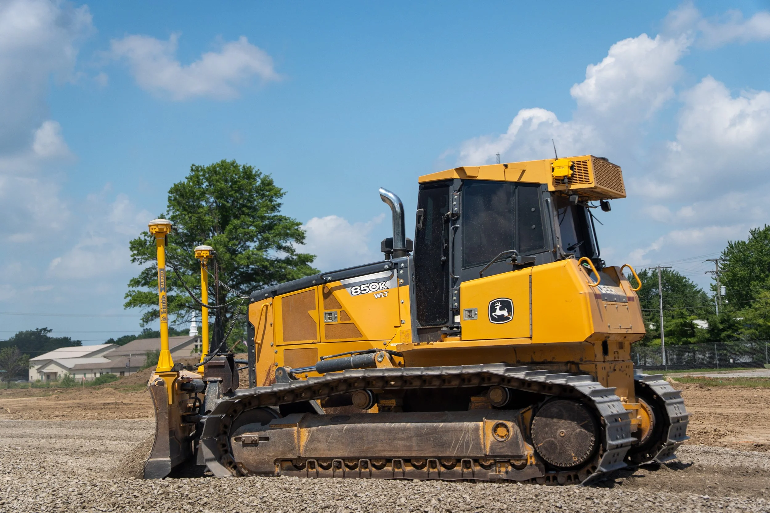 A yellow John Deere bulldozer working on a construction site with a blue sky and scattered clouds, trees, and houses in the background.