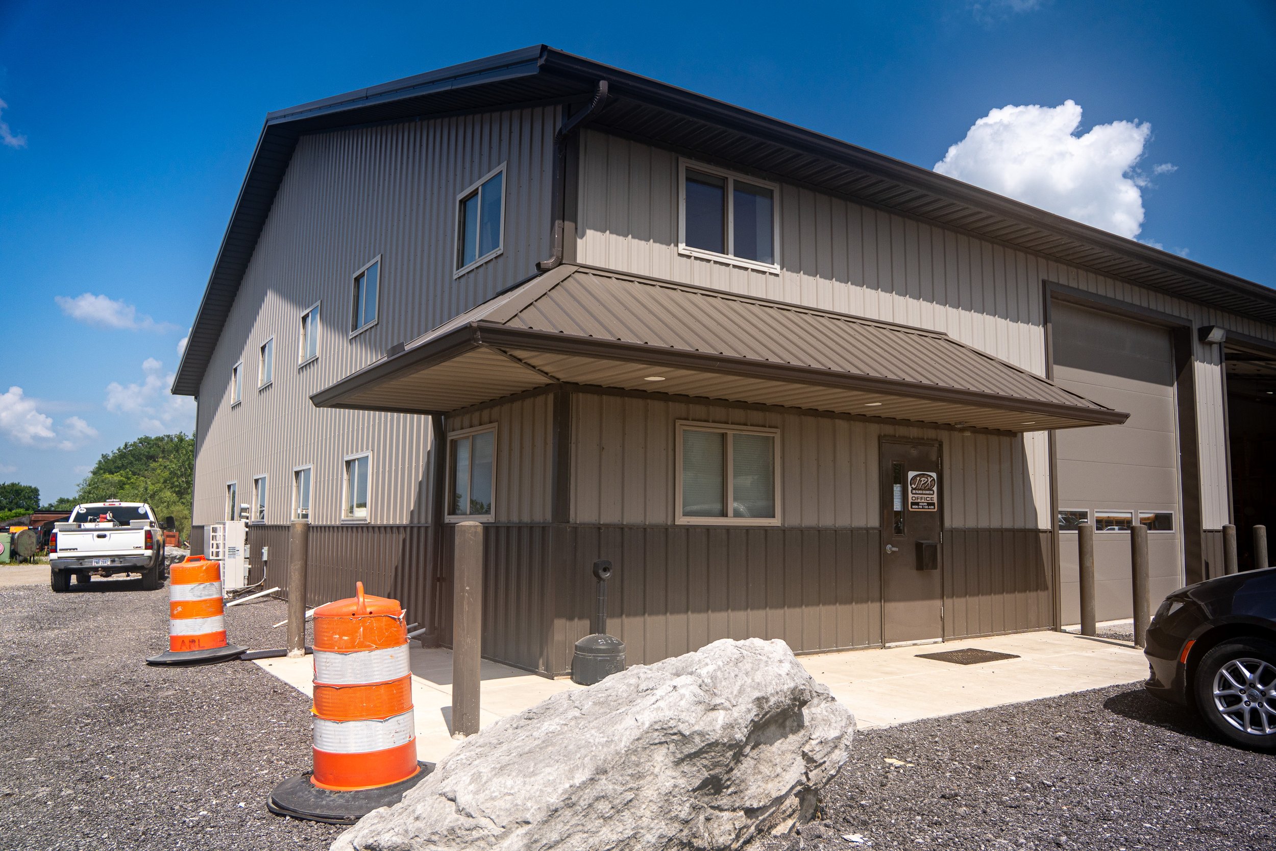 A two-story industrial building with beige metal siding and several windows under a blue sky with clouds. There are construction cones, a large rock, and parked cars in the foreground.