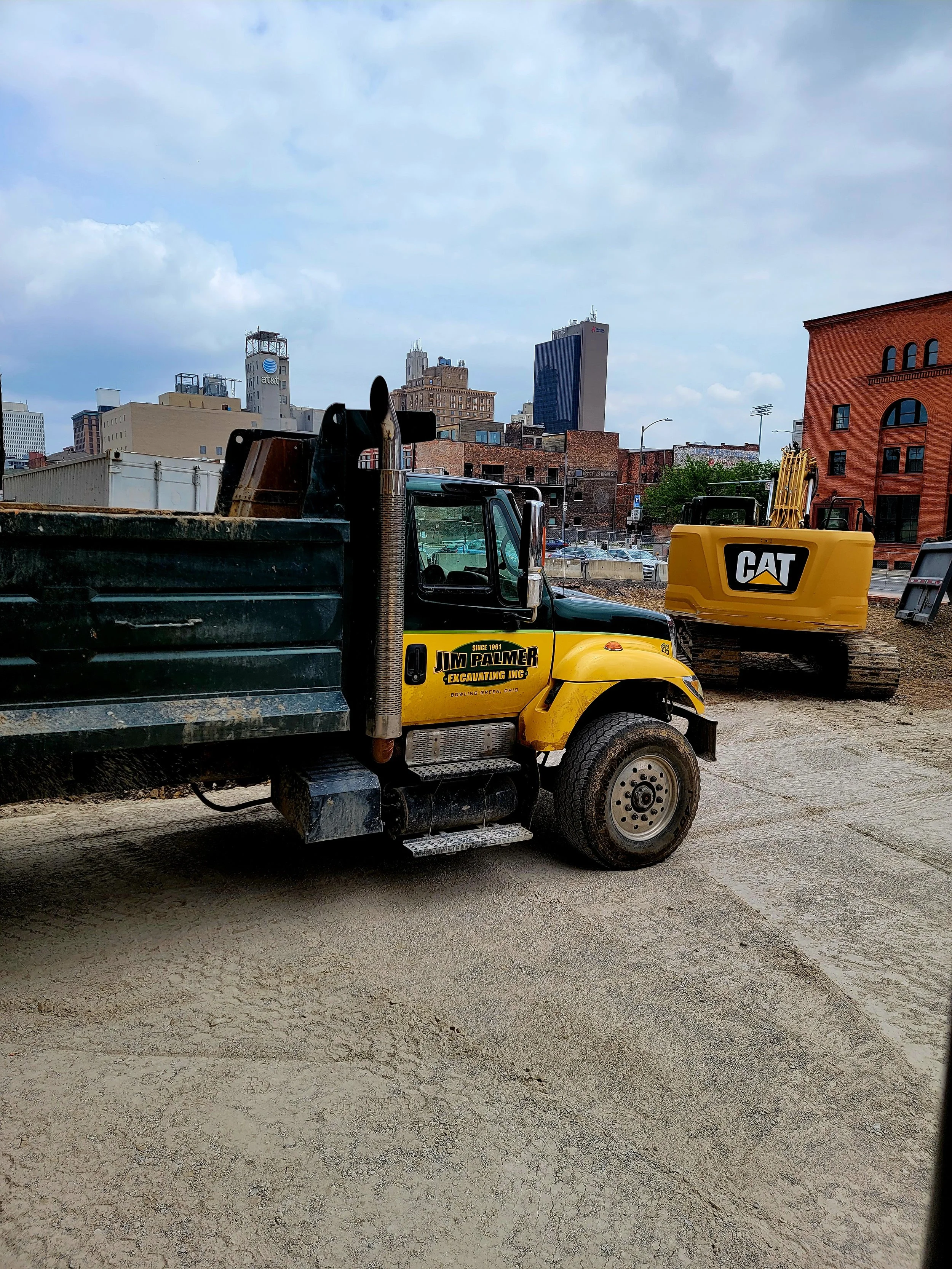 Construction site with a yellow CAT excavator and a black dump truck, with a city skyline in the background under a cloudy sky.