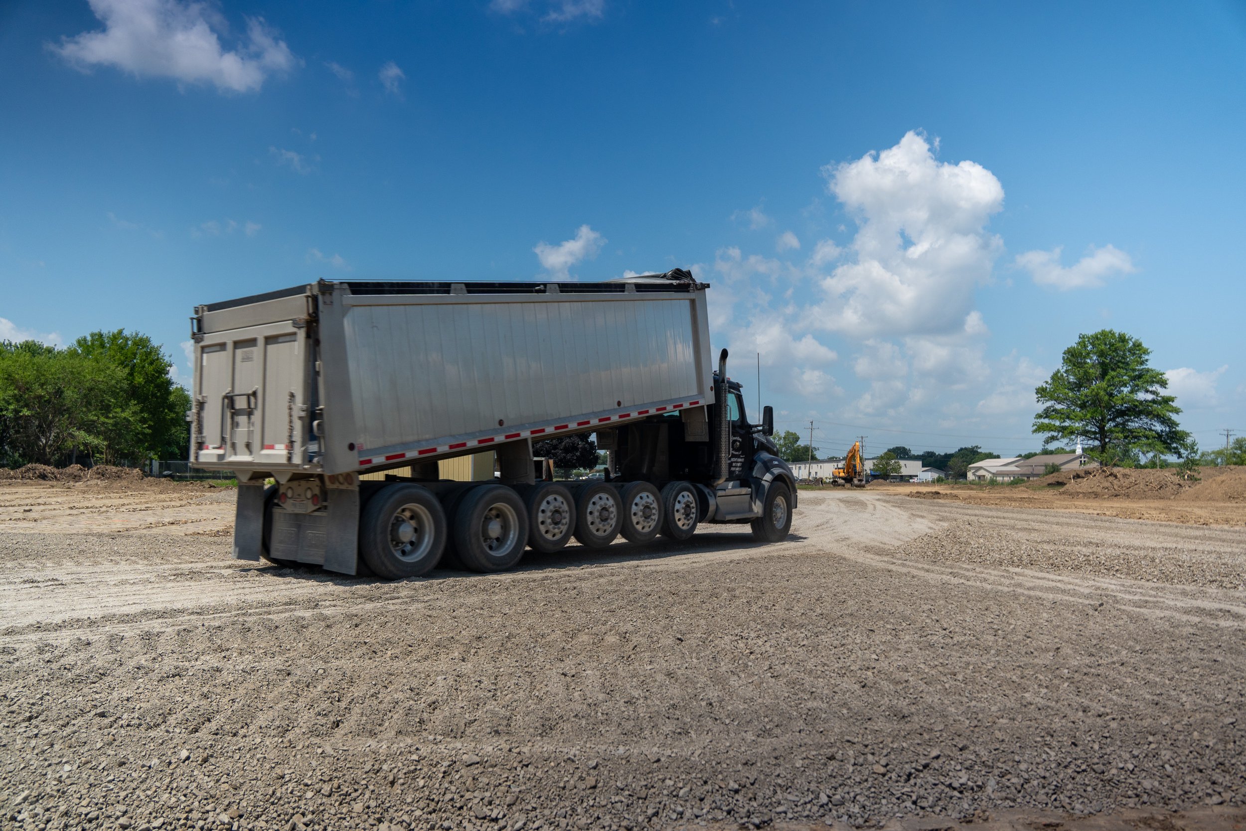 A construction site with a truck, dirt ground, trees, and a blue sky with scattered clouds.
