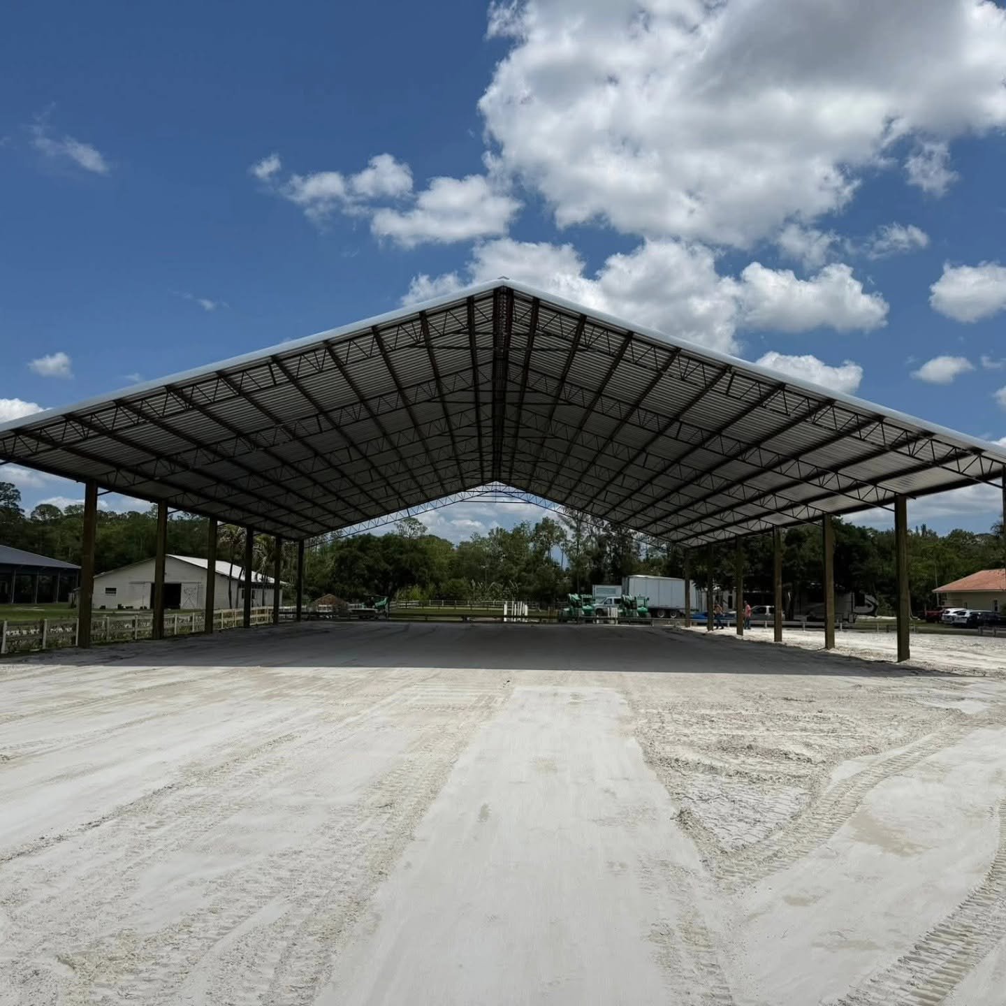 A large open-sided metal carport structure with a dirt ground beneath it, set against a blue sky with scattered clouds. There are trees, small buildings, and trucks in the background.