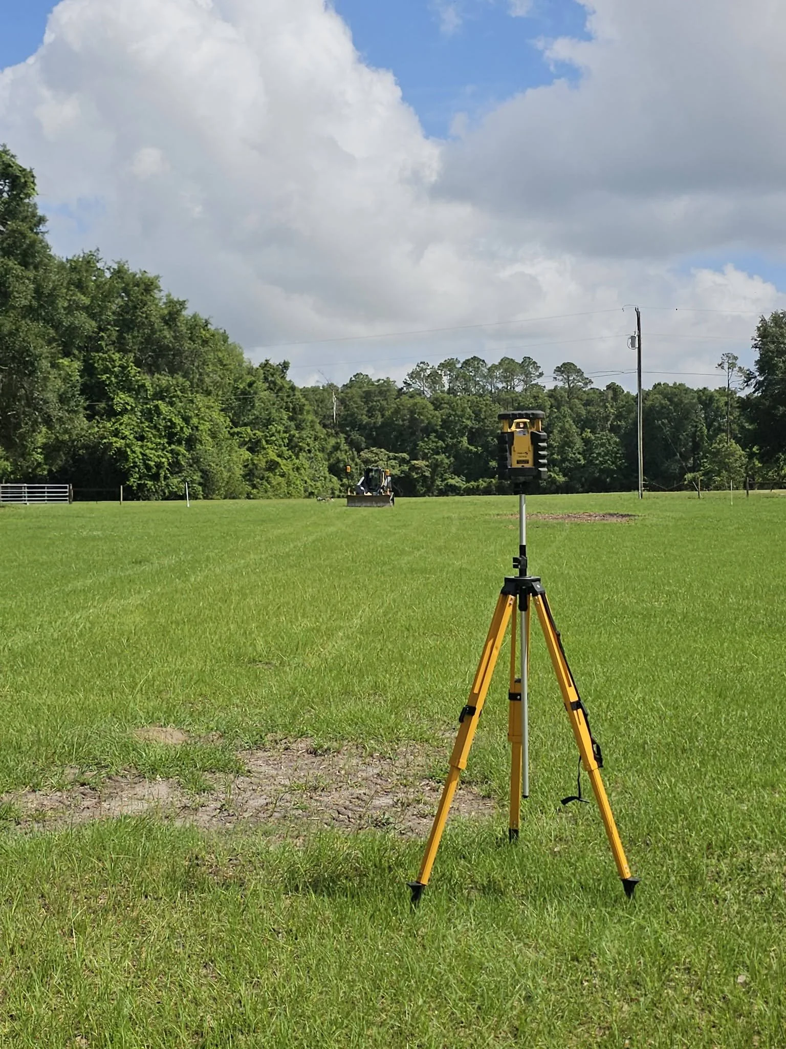 A surveyor's tripod with a mounted electronic device on a grassy field, with a backhoe in the background, trees along the horizon, and a partly cloudy sky.