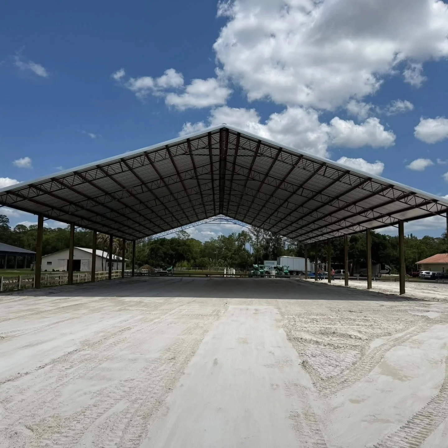 Large open-sided metal-roof pavilion under a blue sky with clouds, situated on a dirt ground with tire tracks, surrounded by trees and rural buildings.