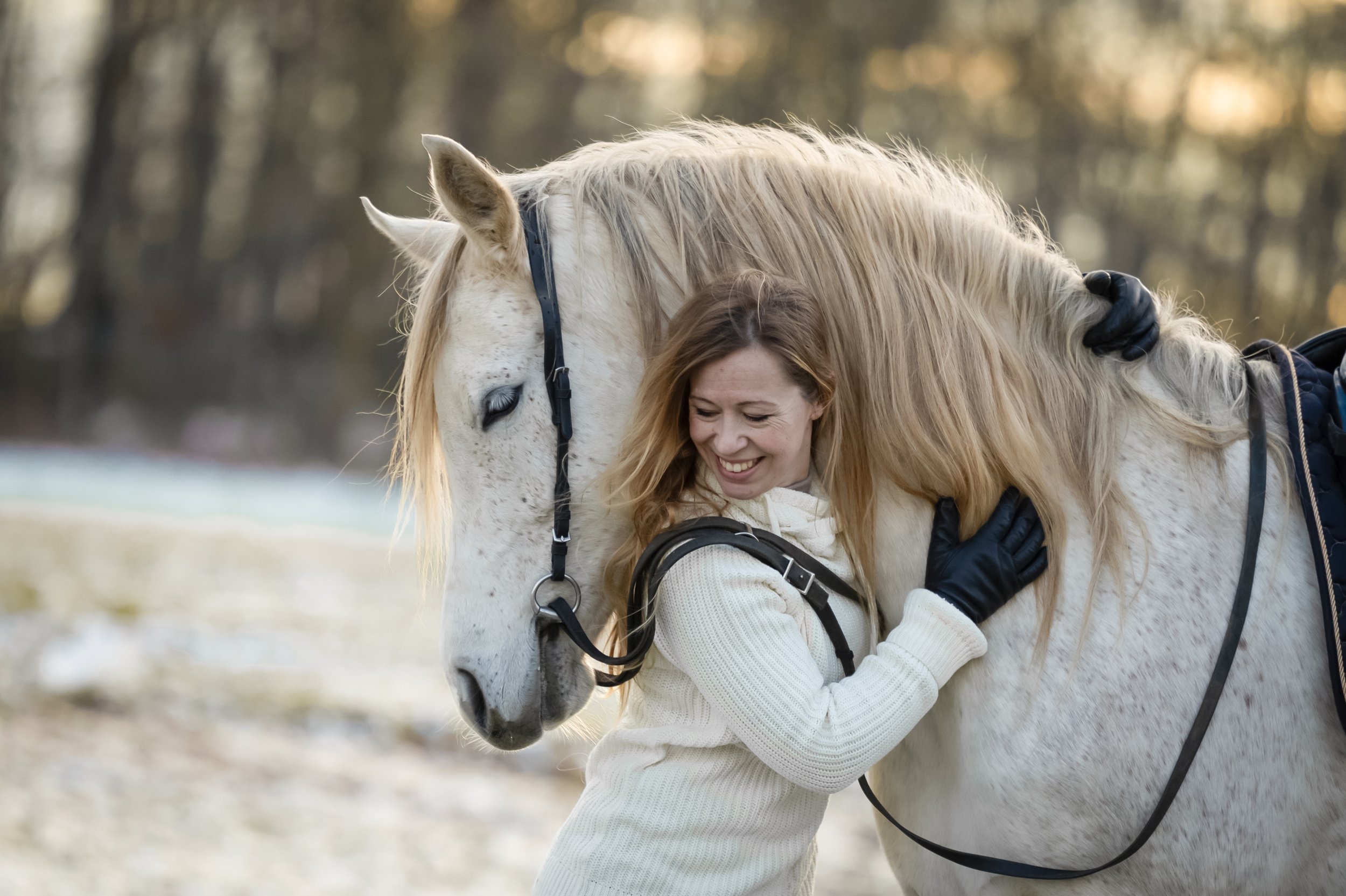 Eine Frau in einem weißen Pullover umarmt ein weißes Pferd im Freien, während sie lächelt.