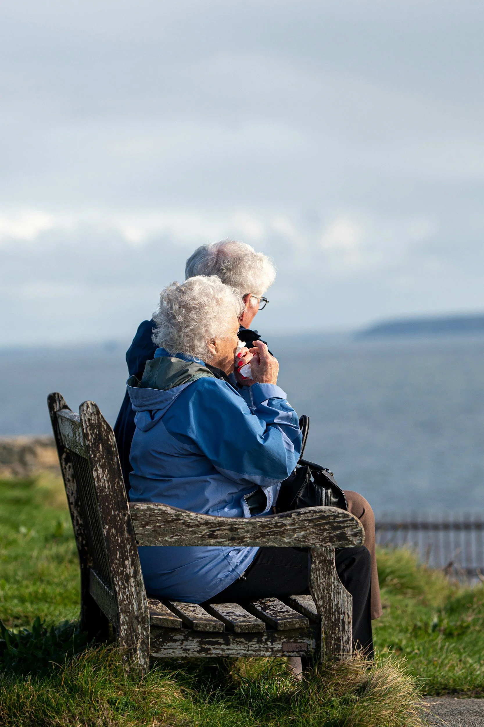 Two elderly women with curly white hair sitting on a wooden bench outdoors, sharing a moment of affection, with a cloudy sky and distant sea in the background.