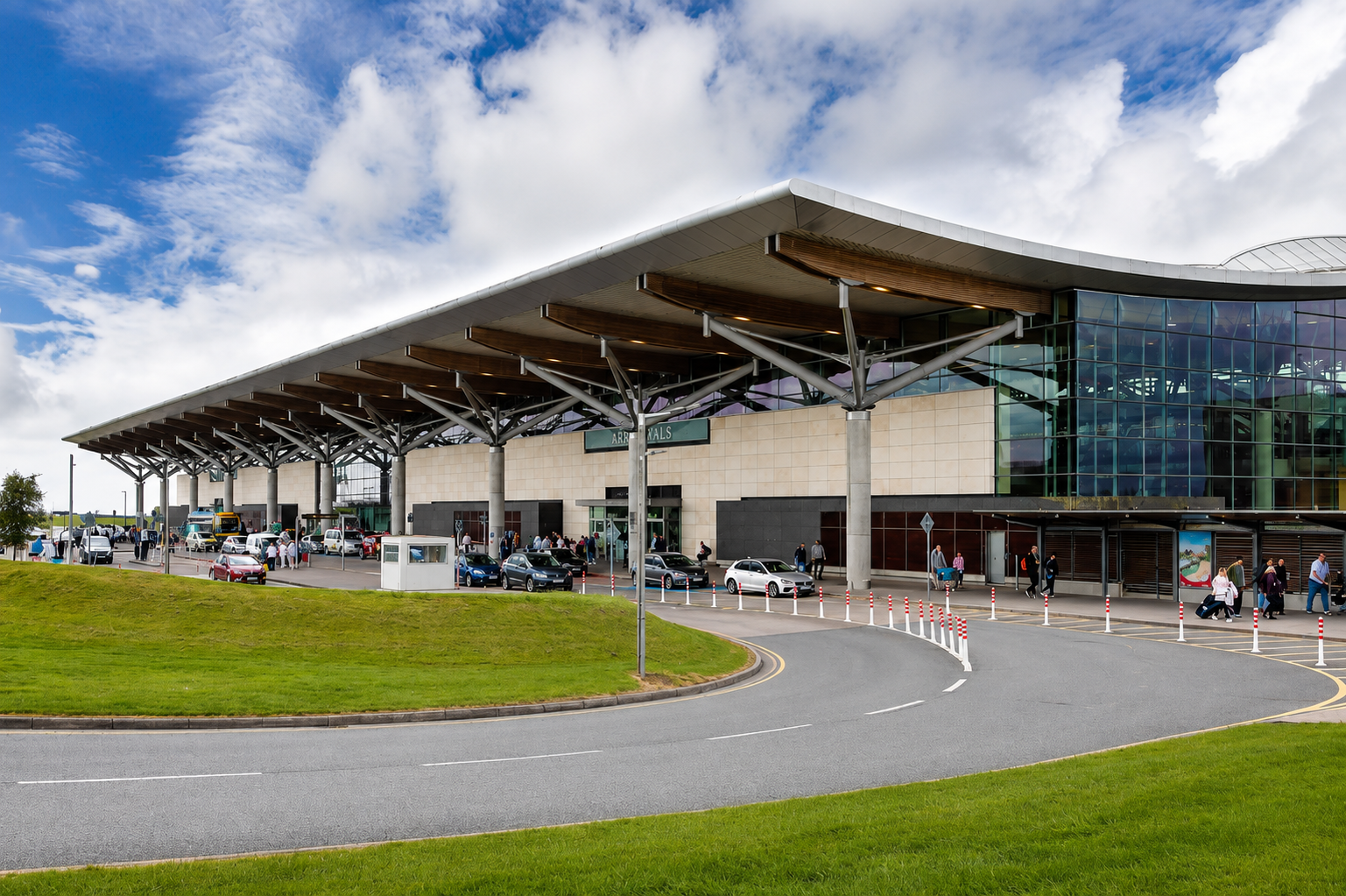 Modern airport terminal with glass facade, busy arrival area with cars and pedestrians, surrounded by green grass, under a partly cloudy sky.