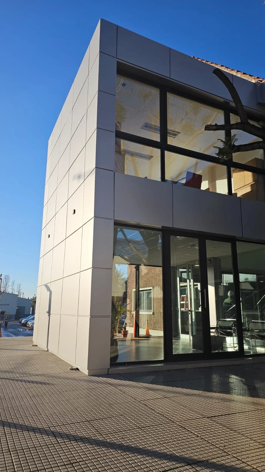 Modern two-story building with large glass windows and a white exterior, set against a clear blue sky. The ground is paved, and there are some orange traffic cones inside the building near the entrance.